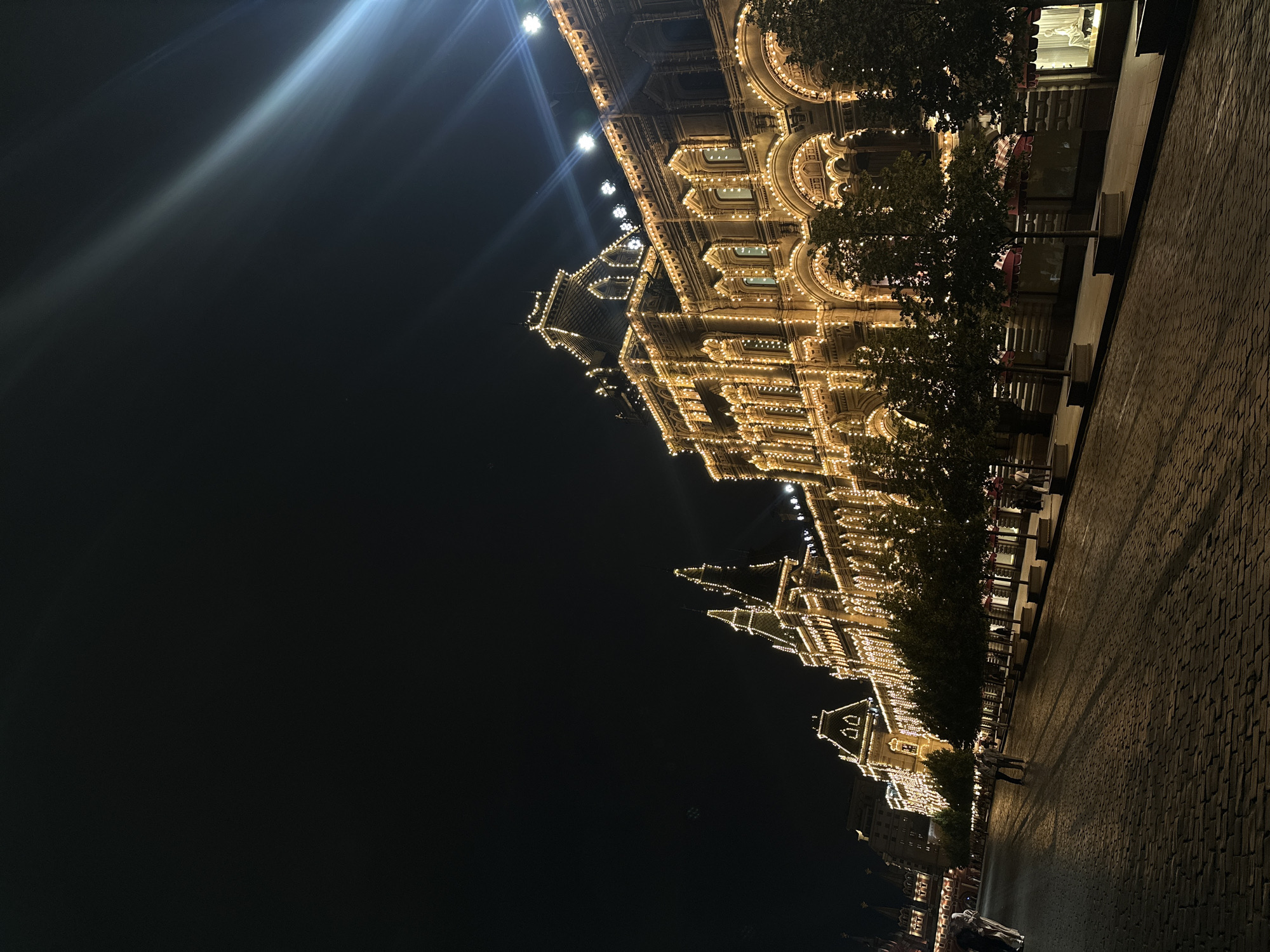 This image depicts Red Square in Moscow, Russia at night. A grand, historic building, illuminated by countless warm-toned lights, dominates the right side of the frame. The building features intricate architectural details, including arched windows and ornate rooflines. Rows of young trees with lush green foliage line the facade of the building, casting long shadows across the cobblestone ground. The cobblestone pavement stretches into the distance, leading towards other buildings that are also partially illuminated. Several individuals are visible in the scene, appearing as silhouettes or indistinct figures walking along the square and the sidewalk. One person is seen walking towards the foreground on the left, while others are more spread out further down the square. The sky above is dark, with a bright light source, possibly a spotlight or a distant reflection, creating a lens flare effect in the upper right corner. The overall impression is one of a beautifully lit urban landscape during the evening.