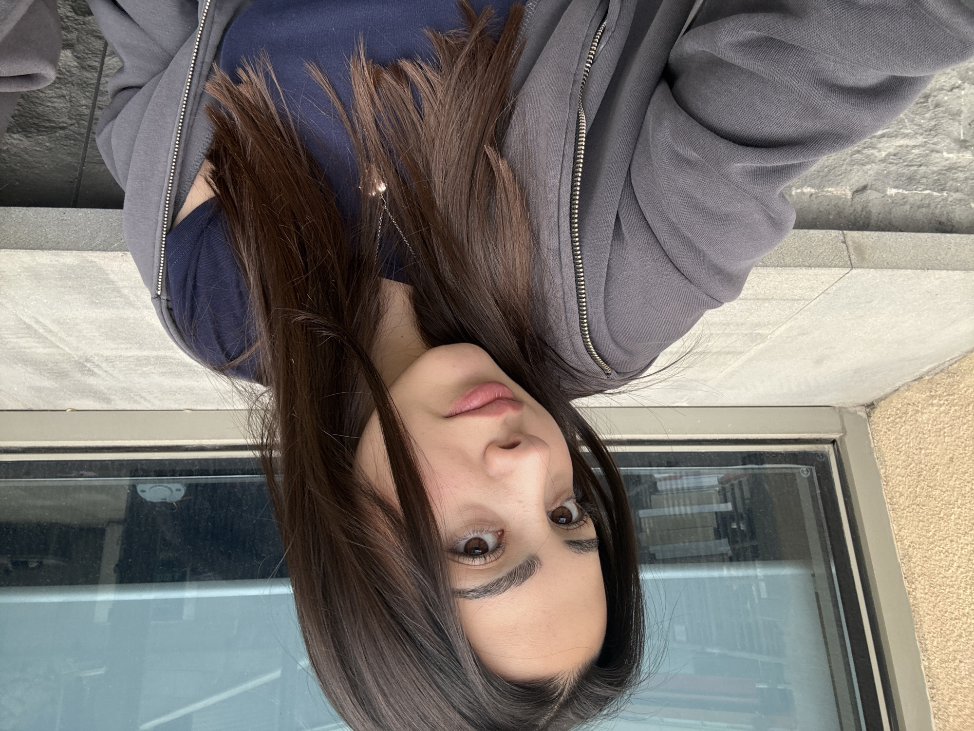 A woman is indoors with her face and upper body visible. She has long, dark brown hair that partially obscures her face. She is wearing a dark blue shirt under a gray zip-up hoodie. A delicate silver necklace is visible on her chest. Her skin appears fair. She looks directly at the camera with dark brown eyes.
Behind her is a window that appears to be a ground-level or basement window, judging by the proximity of the ground and what appears to be a wall or ledge outside. The window reflects the interior, showing a blurry image of books. The color and texture suggest that the external environment is outdoors, possibly with stone or concrete surfaces.
It is not possible to determine the location in Yerevan, Armenia from this image.