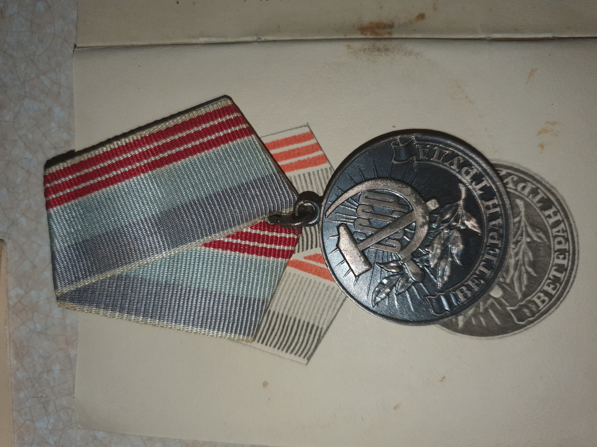 A medal with a hammer and sickle emblem hangs from a ribbon with red and grey stripes. The medal is circular and dark in color, with raised text around the rim. The ribbon is attached to a loop on the top of the medal. The medal appears to be resting on a light-colored, textured surface, possibly paper or fabric. The background is out of focus. There is a faint shadow cast by the medal.