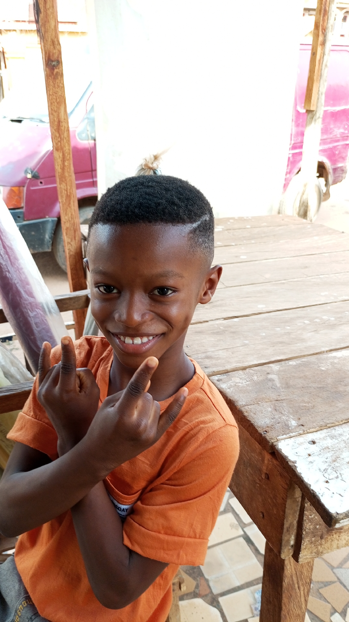 A young Black boy, approximately 8-12 years old, is depicted smiling broadly at the camera, showcasing dimples on his cheeks. He has a short, dark haircut with a shaved line design visible on the right side of his head. He wears a bright orange short-sleeved t-shirt and grey shorts. With his arms crossed over his chest, he holds up both hands, forming a "rock on" or "devil horns" gesture.
The setting appears to be outdoors or a semi-open space, likely during the daytime, indicated by the bright, even lighting. To the boy's right is a rustic wooden table, constructed from planks, showing signs of wear with chipped white paint. In the background, a tall wooden pole stands vertically. Further back, parts of two reddish-maroon vehicles are visible, one on the upper left with a dark bumper, and another on the upper right. A light-colored wall or structure is also visible behind the boy. A piece of fabric or plastic is tied to a pole in the mid-background. The ground features an assortment of light-colored, possibly broken, tiles. The boy's expression is joyful and playful. A small white tag with illegible blue text can be seen on the left sleeve of his t-shirt. The scene conveys a relaxed and happy moment in an outdoor environment in Sabon Tasha, Nigeria.