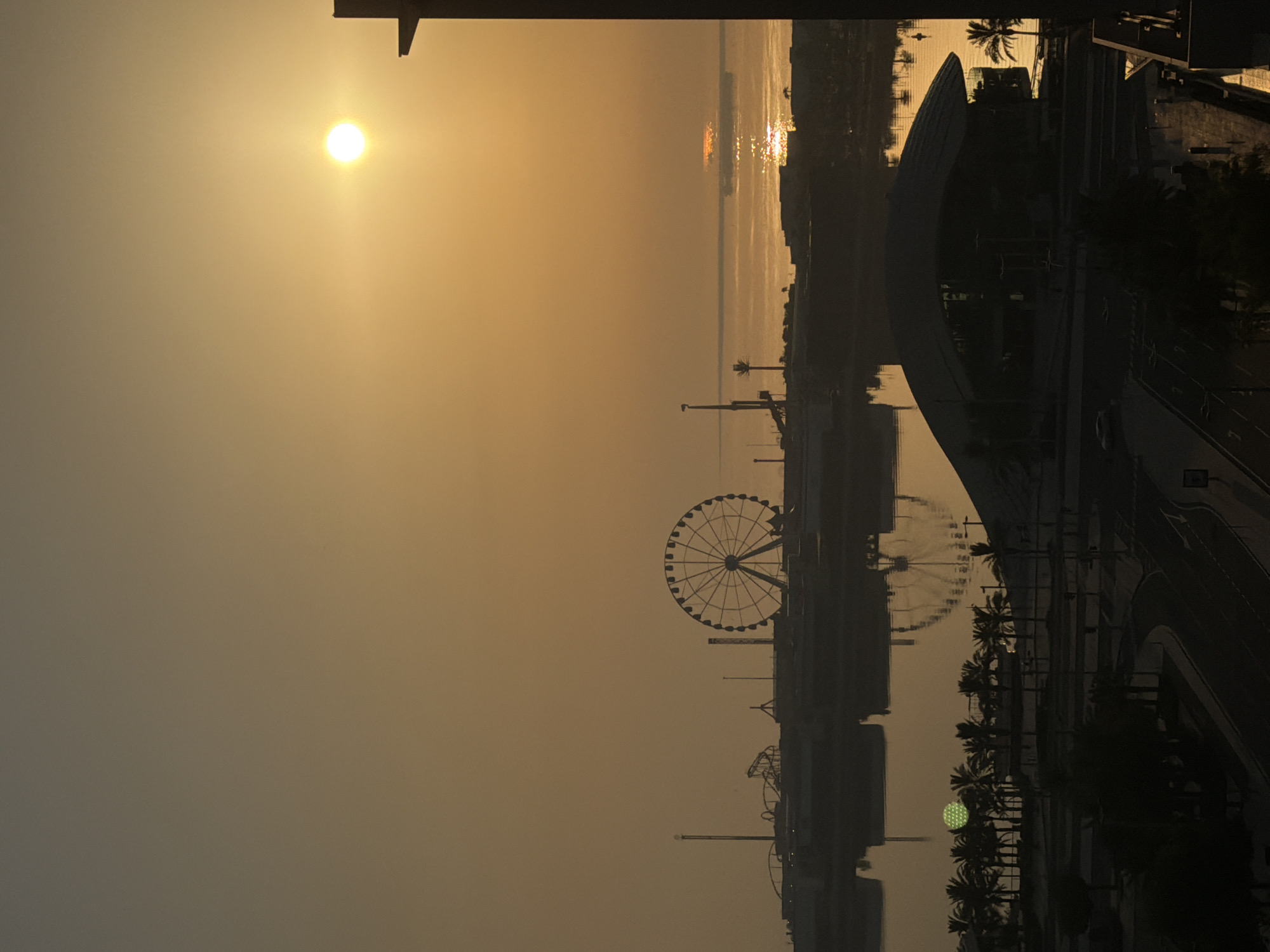 A silhouette of a Ferris wheel and roller coaster track is visible against a hazy, golden sky during sunrise. The sun, a bright orb, is positioned high in the sky, casting a warm glow over the scene and reflecting on the water. In the foreground, palm trees and a modern building are silhouetted, with a road and a few cars visible. The water mirrors the Ferris wheel and the sky, creating a symmetrical effect. The overall impression is one of a tranquil morning in a coastal city.