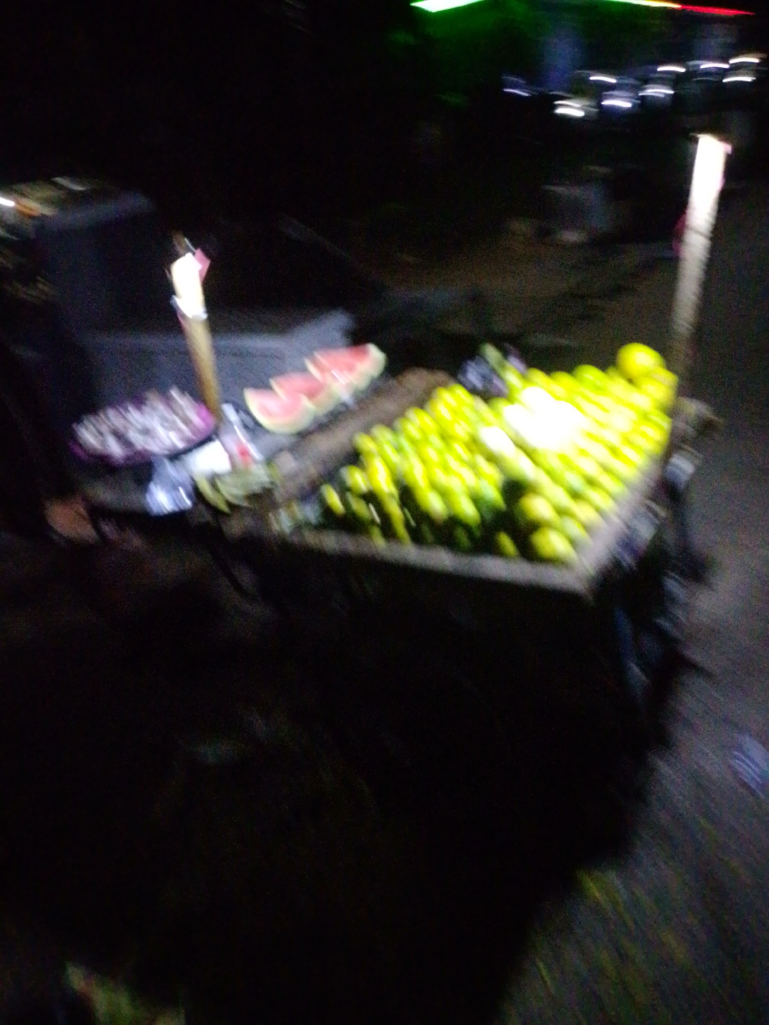 In Jalingo, Nigeria, a blurry, low-light image captures a street-side fruit stall at night. A wooden cart is laden with a large quantity of green, round fruits, likely limes or lemons, arranged in neat rows and illuminated by a bright light. To the left of the cart, slices of watermelon are visible on a surface, accompanied by what appears to be a pile of packaged items, possibly snacks or other goods. A vertical pole with a light fixture stands near the watermelon, and another pole with a similar light is visible further to the right. The background is dark and indistinct, suggesting an outdoor urban setting with some distant, out-of-focus lights and colors. The overall impression is of a bustling night market scene.
