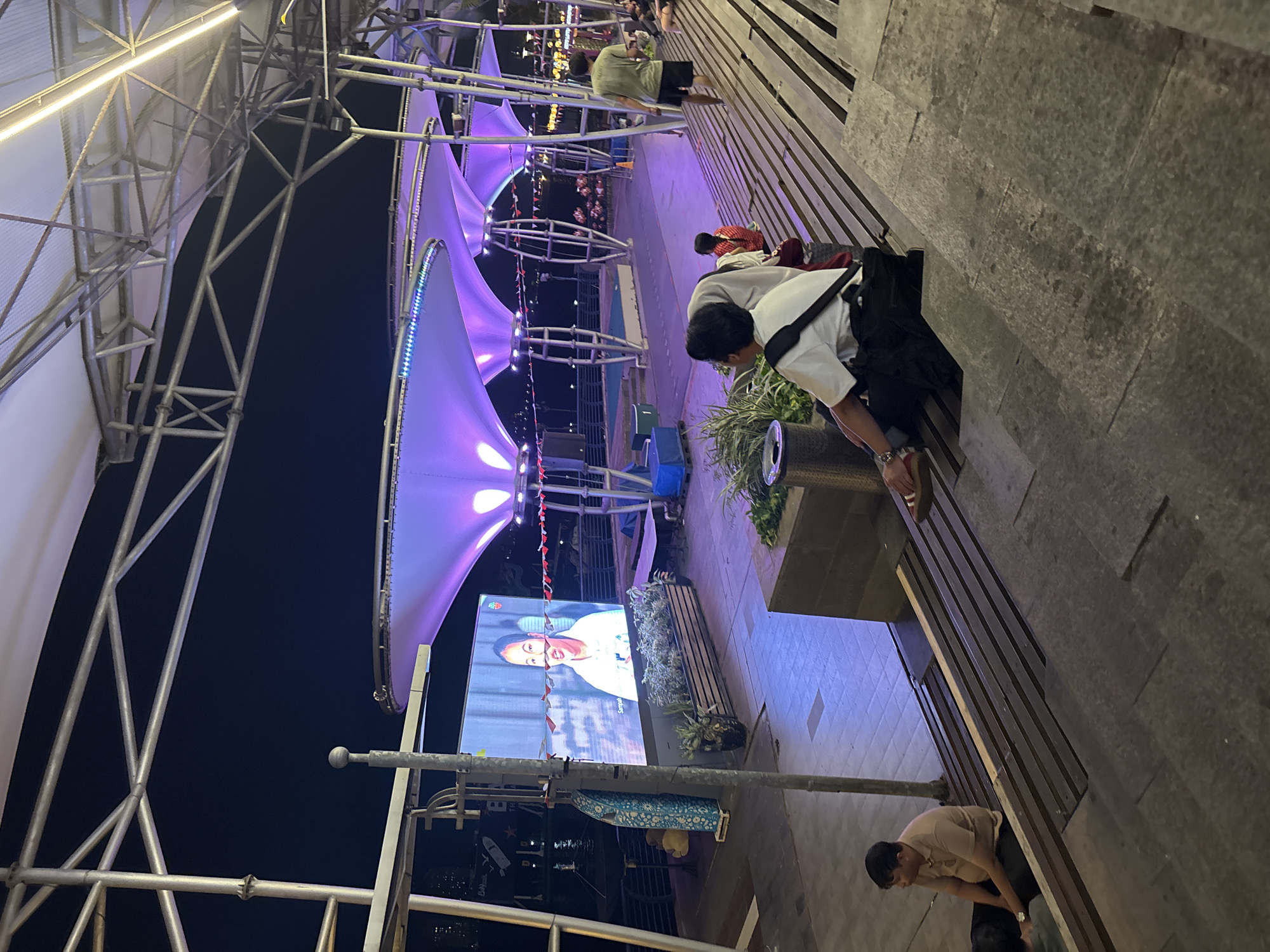 The image depicts an outdoor nighttime scene at a waterfront entertainment area in North Jakarta, Indonesia. A large outdoor screen displays a close-up of a woman speaking. In the foreground, several people are seated on wide, tiered wooden steps. One person in the middle ground is sitting facing away from the camera, wearing a white shirt and black pants, with a black bag slung over their shoulder. To their left, another person is seated and looking towards the screen. Further down the steps, a man in a light brown polo shirt and dark pants sits facing left. In the background, people are walking and sitting, and a man in a green shirt and black shorts walks up the steps. The area is covered by a large, illuminated canopy with purple lighting. The waterfront can be seen in the distance, with lights from buildings and boats reflecting on the water.