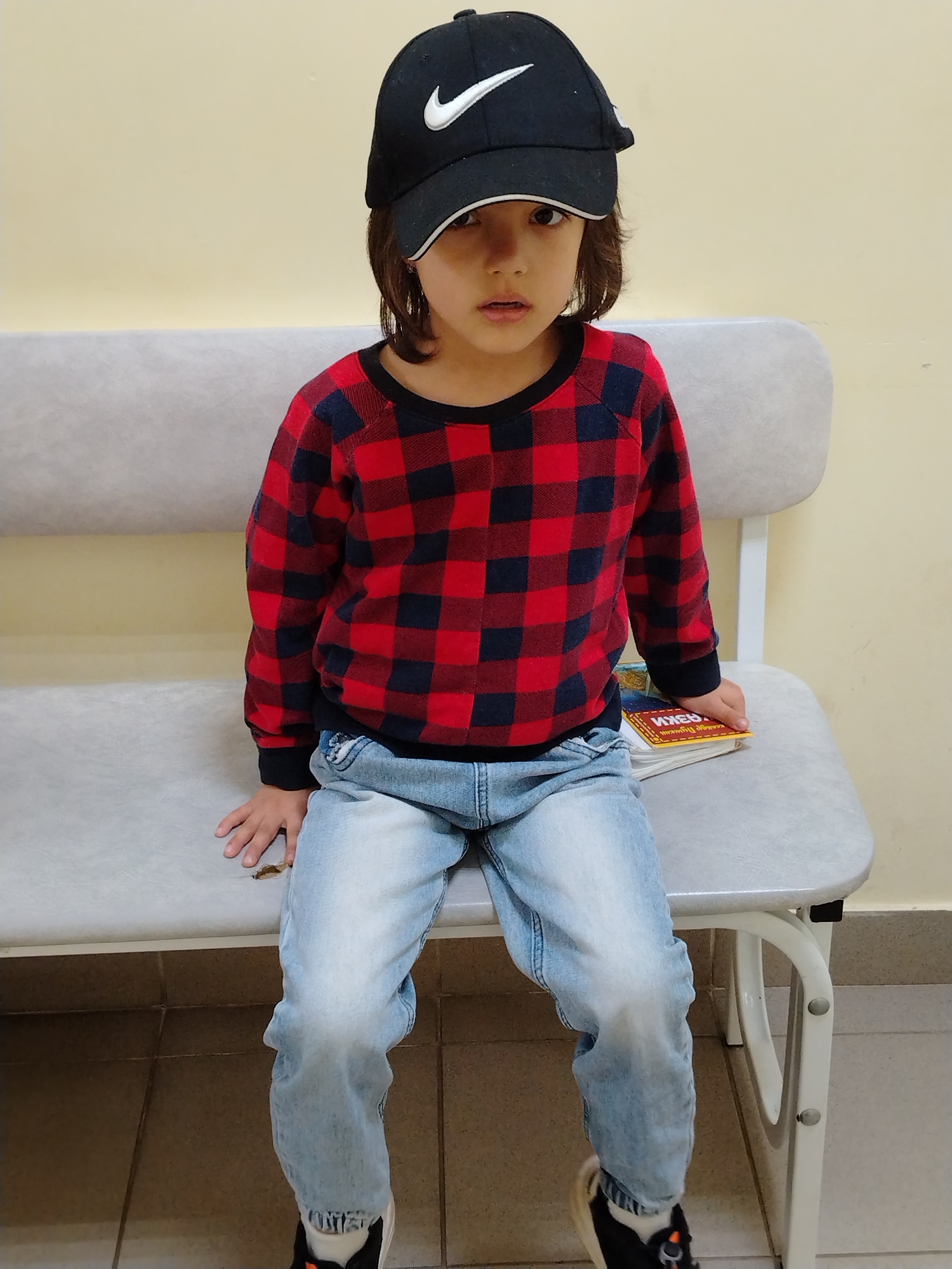 A young child with dark hair, wearing a black Nike baseball cap, a red and blue plaid long-sleeved shirt, and light-wash jeans, is seated on a gray bench. The child is looking directly at the camera with a neutral expression. To the child's right, a stack of books with a yellow cover is visible. The background consists of a plain, light-colored wall and a tiled floor. The lighting appears to be artificial, originating from above. The scene is indoors and suggests a waiting area or a public space. The location is identified as Noviy, Russia.