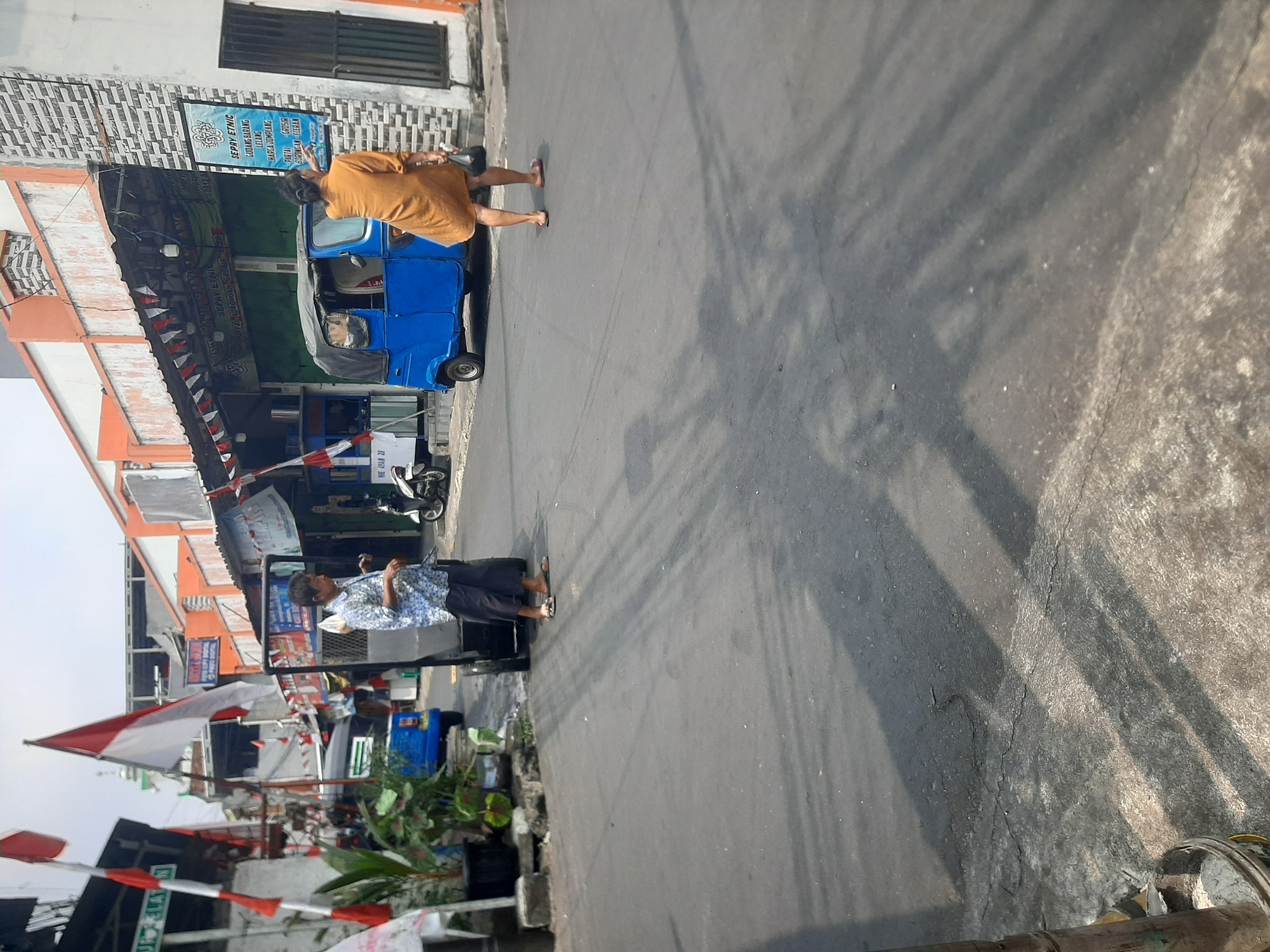 In Central Jakarta, Indonesia, an outdoor street scene unfolds under a bright sky. The asphalt road dominates the foreground, marked with faint tire tracks and dappled with distinct shadow patterns. To the left, a black trash receptacle sits next to a pole.
Further back, on the street, a man in a blue patterned shirt and dark pants stands near a wheeled cart. He is facing away from the camera, appearing to be engaged with something out of frame. To his right, a bright blue three-wheeled vehicle, a common mode of transport in Southeast Asia, is parked. A woman in a long, mustard-colored dress is seen walking away from the blue vehicle, carrying a dark bag.
The background reveals a row of buildings with orange and grey facades, some adorned with festive red and white banners, suggesting a possible celebration or event. Several motorcycles are parked along the roadside. One building displays a large blue sign with text in Indonesian, indicating a business. The overall impression is of a bustling, everyday street scene in an Indonesian city.