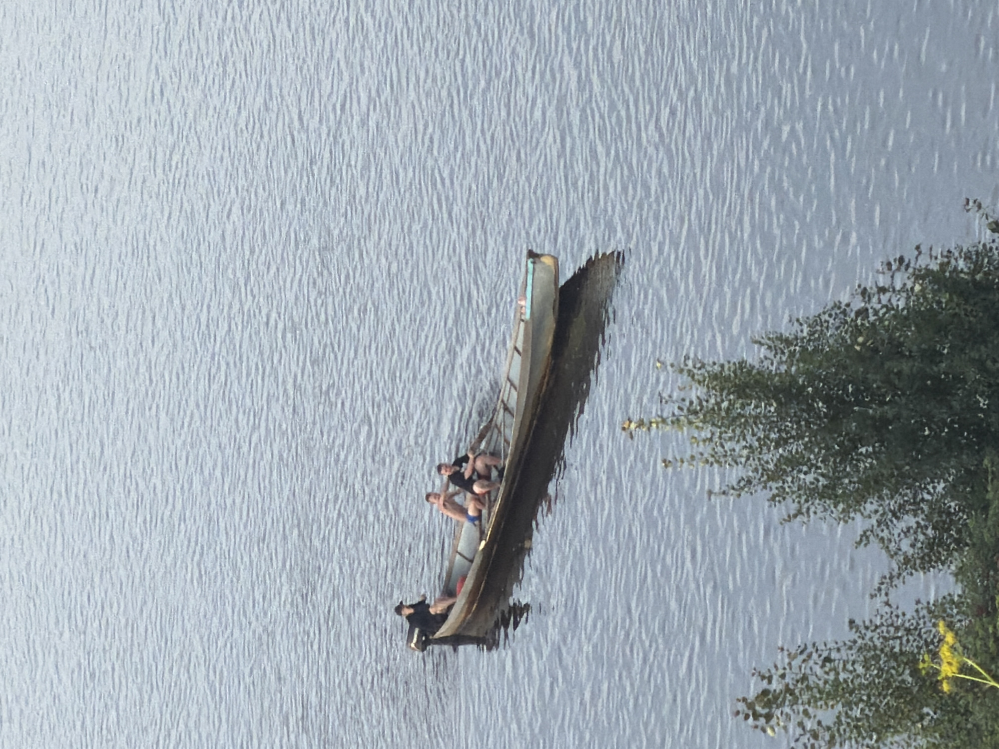 In the city of Cherdyn, Russia, a long, narrow wooden boat with a small outboard motor is on the water. Four individuals are in the boat. One person, wearing a black shirt and a dark cap, is seated at the stern, appearing to operate the motor. Three other individuals are seated in the middle and forward sections of the boat. One person in the middle is wearing blue shorts and appears to be looking towards the right. The other two individuals are wearing dark shirts and are seated closer together. The water surface is rippled, suggesting a gentle breeze. In the foreground, a portion of a tree with green foliage and a few yellow wildflowers is visible. The overall scene depicts a leisurely activity on the water.