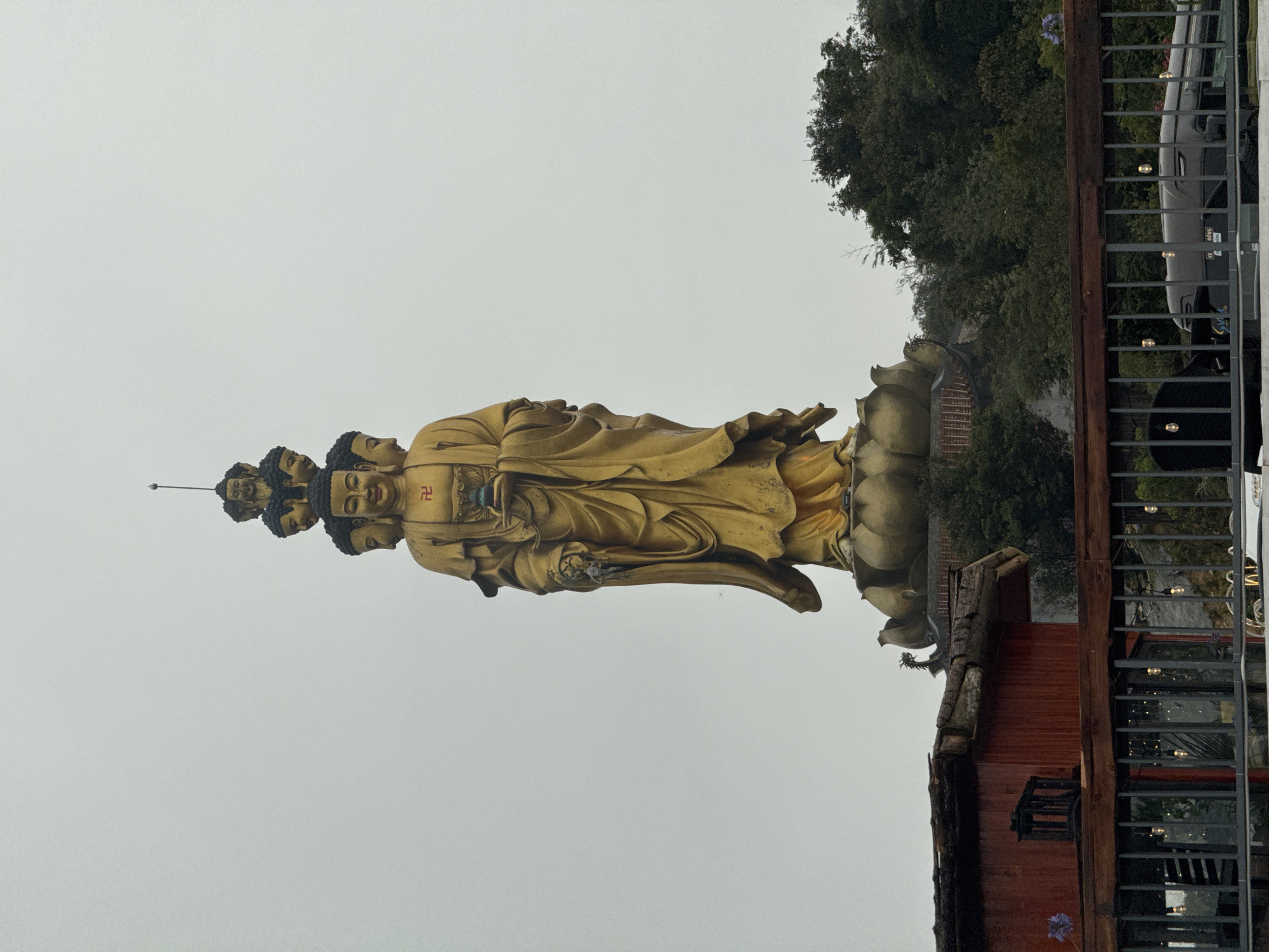 A tall, golden statue of a Buddhist figure with multiple heads, standing on a lotus-like pedestal, dominates the image. The statue is located outdoors, in Binh Lu, Vietnam. The background features a muted, overcast sky. A tree line and foliage are partially visible behind and to the right of the statue. A small building with a rustic roof is situated at the lower right of the statue. A fence with decorative lighting separates the statue from the building and the surroundings.