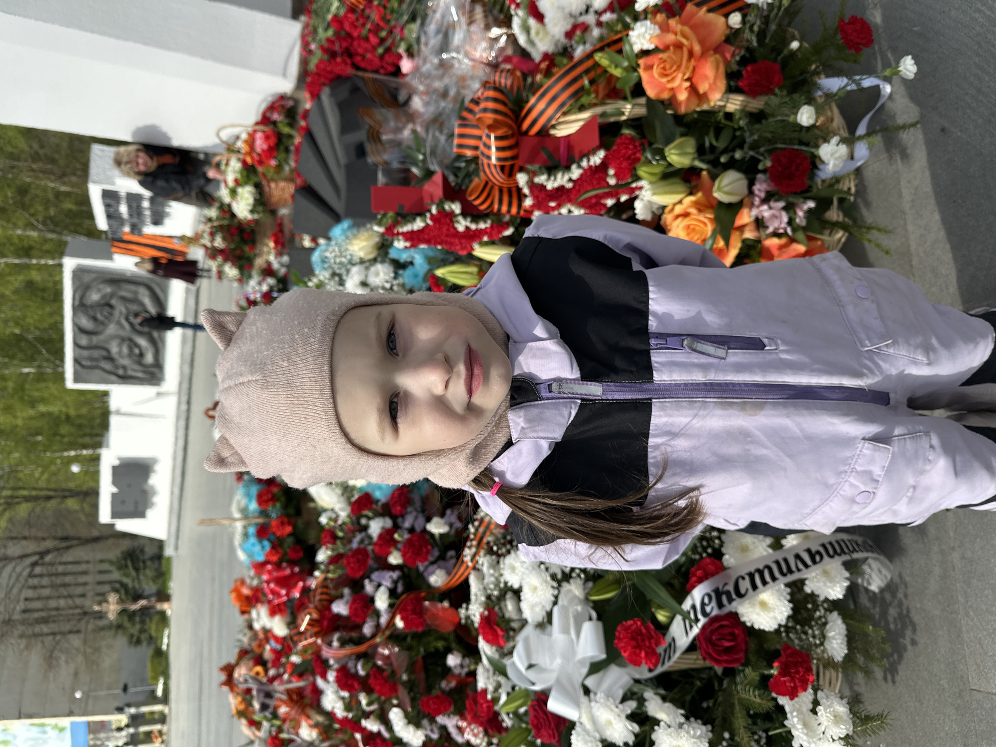 A young girl stands among floral wreaths in a public space. The child wears a light purple jacket with black accents and a tan cap with small ears. A ponytail is visible. The wreaths are composed of red, white, orange, and blue flowers, along with ribbons, including a St. George ribbon. In the background is a stone monument with relief carvings. People can be seen near the monument. The location is Chaikovsky, Russia.