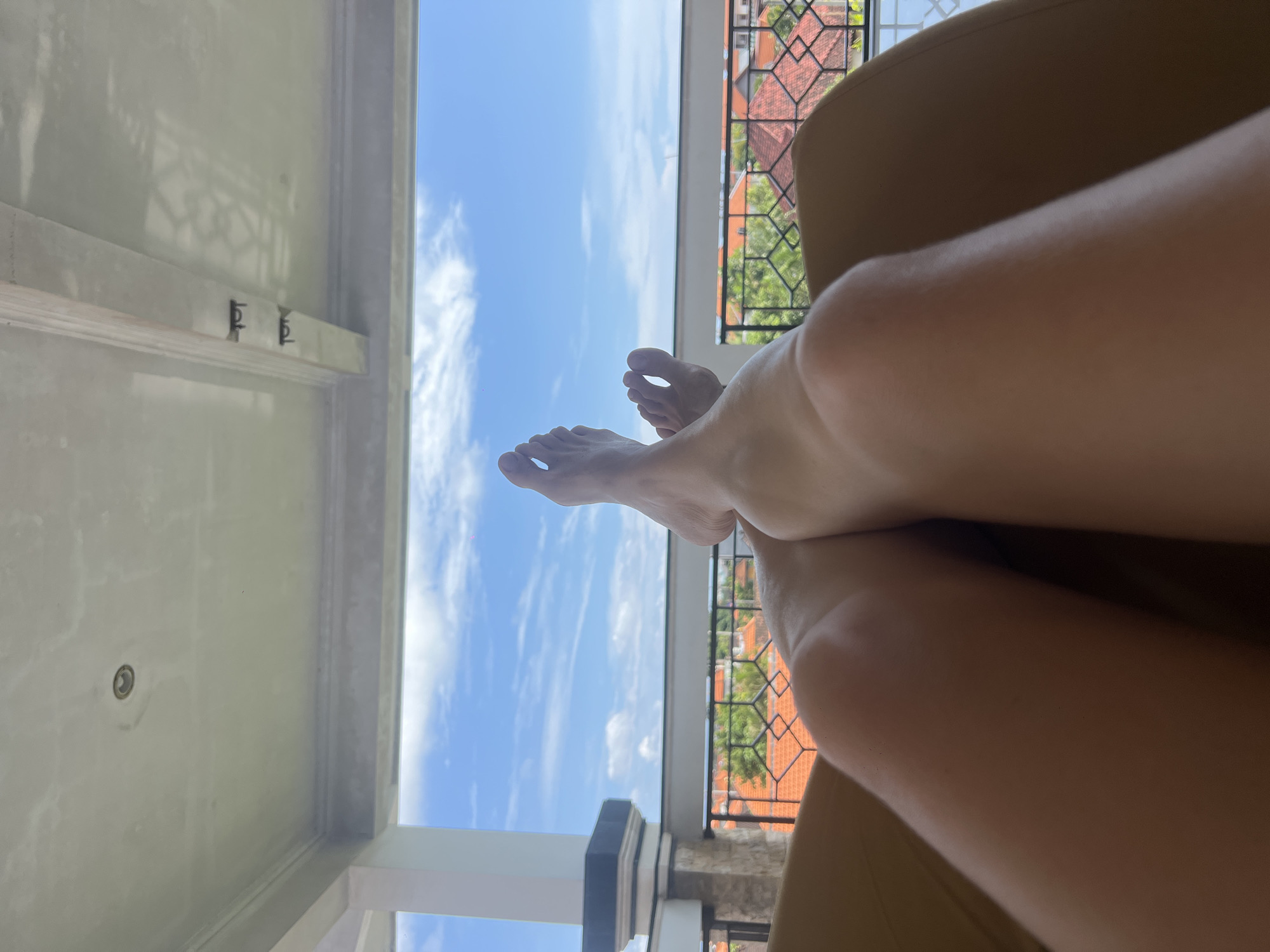 A low-angle shot shows a person lounging in a chair on a balcony. The legs are propped up, feet pointing toward a blue sky with scattered clouds. To the left, the balcony's ceiling is visible, with details like light fixtures and ornate designs. A railing in the background provides a view of buildings with red tile roofs and lush green trees. The location is likely Canggu, Indonesia, based on the architectural style and vegetation. The focus is on relaxation and the view, capturing a moment of leisure.