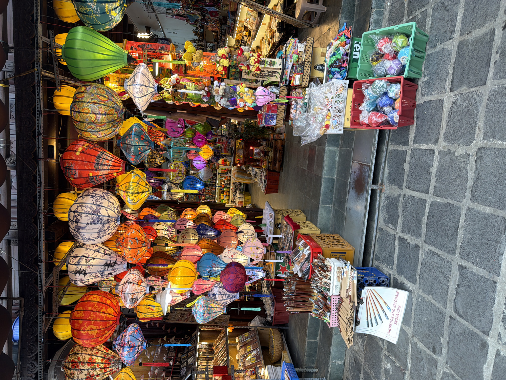The image captures a vibrant street scene in Hoi An, Vietnam, likely a market or shopfront, characterized by a profusion of colorful hanging lanterns. The lanterns, in various shapes, sizes, and patterns, dominate the upper portion of the frame, illuminating the space with a warm glow. They are suspended from the eaves of a building, creating a dense canopy of light and color.
Below the lanterns, the scene unfolds into a bustling marketplace. Stalls are laden with a variety of goods. In the foreground, a display of chopsticks and what appear to be small carved items or keychains is presented on a small stand, with a sign advertising "CHOPSTICK AND KEYCHAIN ENGRAVING." Surrounding this area are baskets filled with more merchandise, including what seem to be small decorative items and perhaps packaged food or sweets.
Further back, shelves and tables display an array of products, ranging from what look like traditional Vietnamese crafts, such as conical hats or small wooden carvings, to more general market goods. The overall impression is one of a lively and colorful commercial environment, typical of tourist areas in Hoi An, where the sale of lanterns is a prominent feature. The architecture suggests traditional Vietnamese building styles with wooden beams and intricate carvings on the shopfront. The ground is paved with large, irregular stone tiles. No people are clearly visible in the foreground, but the presence of stalls and merchandise implies commercial activity.