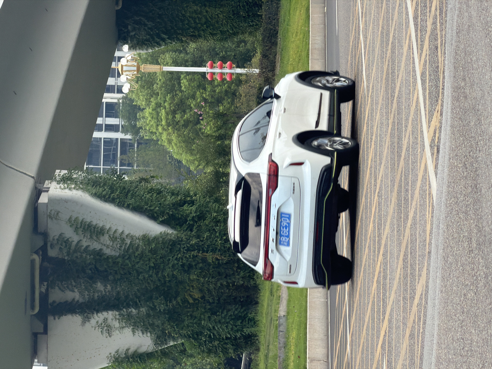 In Jiangjin, China, a white SUV with a license plate reading "8 GE901" is captured in motion on an asphalt road. The vehicle, viewed from its rear quarter, displays a sporty design with a spoiler and neon green accents along its lower body. The road surface is marked with yellow and white crosshatch patterns, indicative of a no-parking zone or a special traffic area. Behind the car, a lush green landscape unfolds, featuring dense trees and a patch of manicured grass. Towering above the scene is a segment of a concrete overpass, its underside partially obscured by climbing green vines. In the middle ground, a decorative streetlamp with multiple bulbs and adorned with red lanterns stands tall, contrasting with the modern architecture of a distant building with reflective windows. The overall environment suggests an urban or suburban setting in Jiangjin, China, with a blend of natural elements and infrastructure.