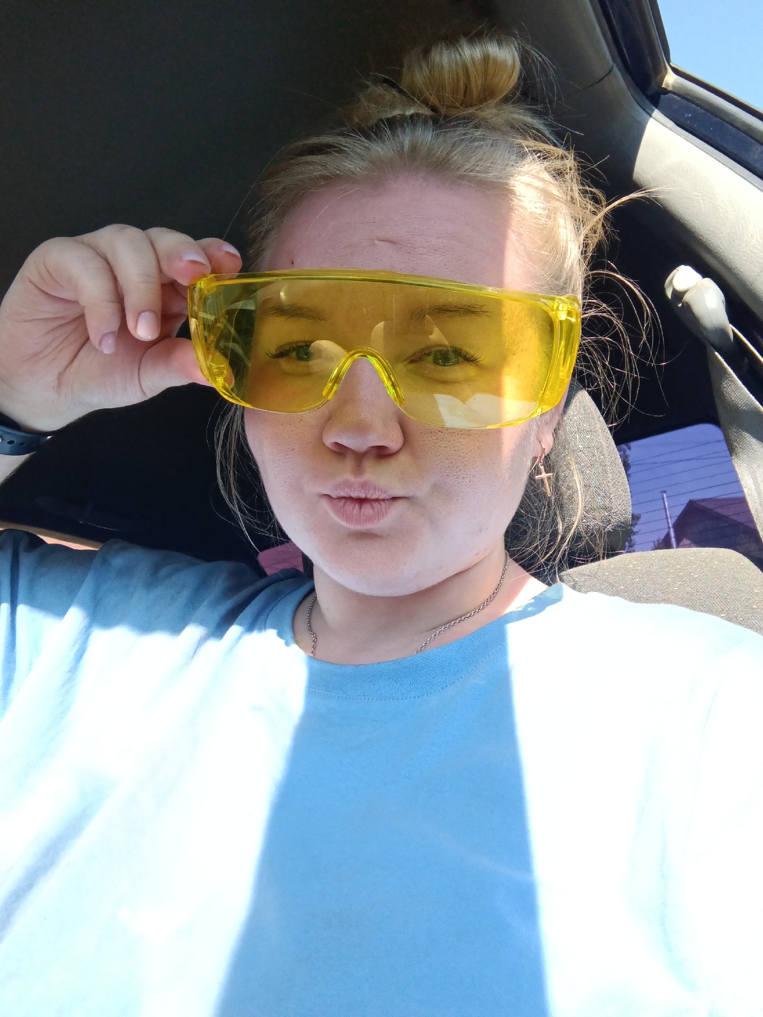 A young woman is seated in a car, wearing a light blue t-shirt and yellow safety glasses. Her blonde hair is styled in a bun. She is holding the glasses with her left hand and pursing her lips in a "duck face" pose. A silver necklace and cross earring are visible. The interior of the car includes a black headrest and a glimpse of the car window and a residential structure outside, implying an urban or suburban setting. There is no explicit indication of the city of Фоки, Russia in the image.