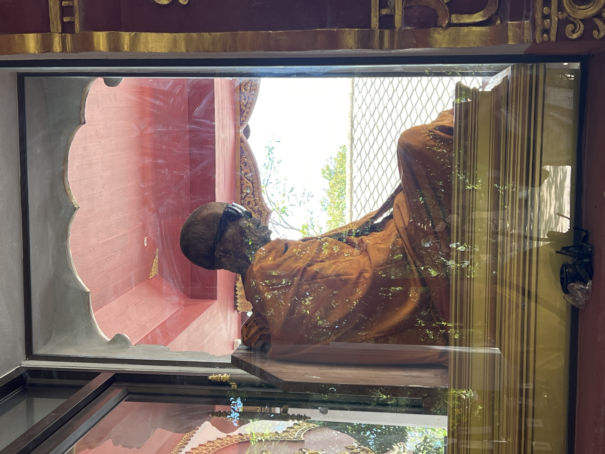 A mummified Buddhist monk wearing sunglasses and an orange robe is displayed in a glass case at Wat Laem Sor Temple in Baan Hua Thanon, Thailand. The monk is seated with his hands resting on his lap. The glass case reflects the surrounding environment, including trees and a temple roof. The monk's body is well-preserved, and his face is visible, though obscured by sunglasses. The robe is adorned with small pieces of gold leaf.