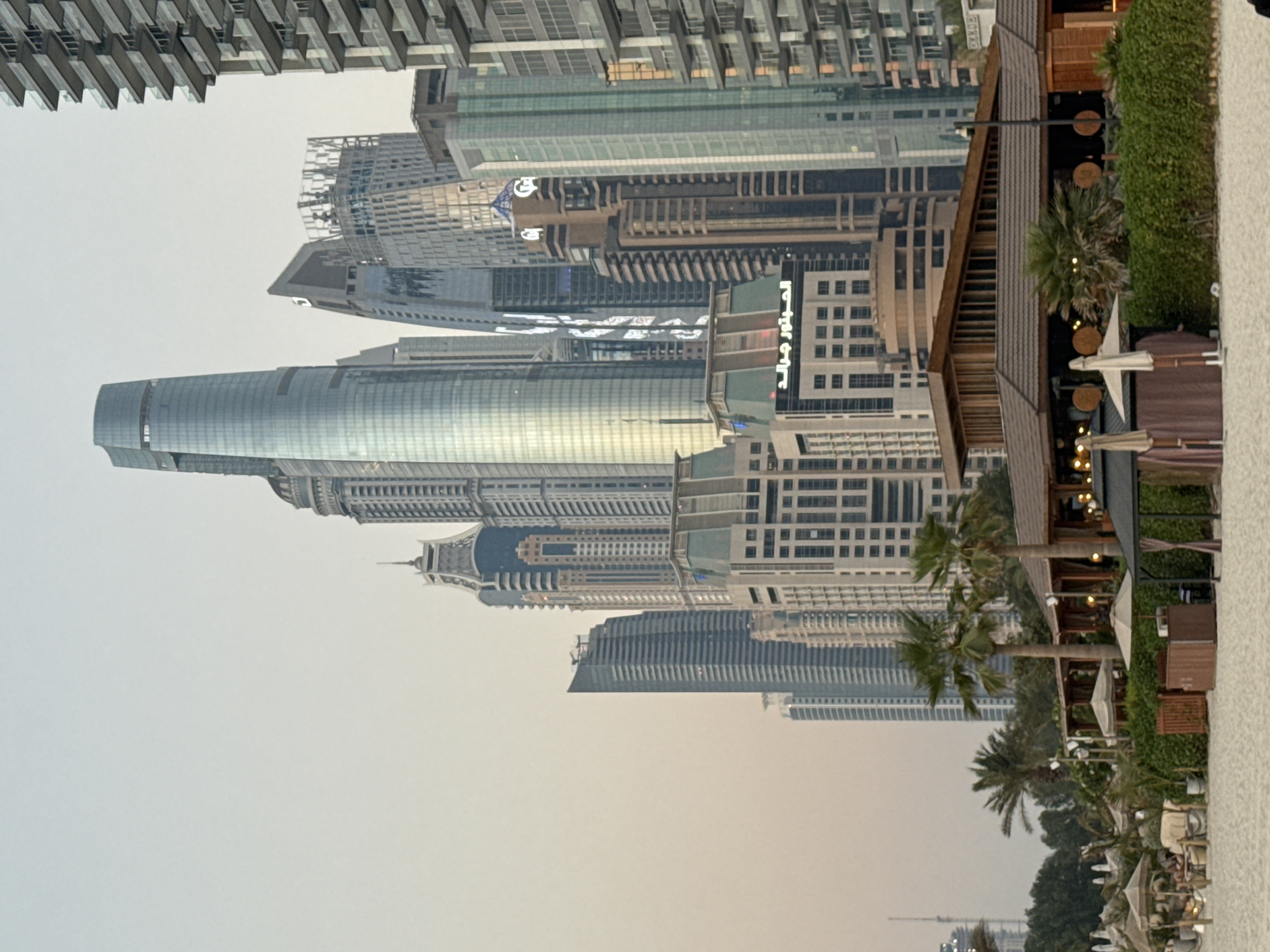 The image captures a serene beach scene in Dubai, United Arab Emirates, with a backdrop of towering skyscrapers. In the foreground, a sandy beach is dotted with umbrellas and cabanas, suggesting a relaxed atmosphere for beachgoers. A low-rise building with a wooden roof, likely a restaurant or beach club, is situated near the water's edge. Palm trees are scattered throughout the scene, adding to the tropical ambiance. Dominating the background is a stunning array of modern skyscrapers, characteristic of Dubai's iconic skyline. Among them, a prominent, slender, glass-clad tower stands out, reflecting the soft, hazy light of the sky. The overall impression is one of a beautiful juxtaposition between natural beach beauty and the architectural marvels of a modern metropolis.
