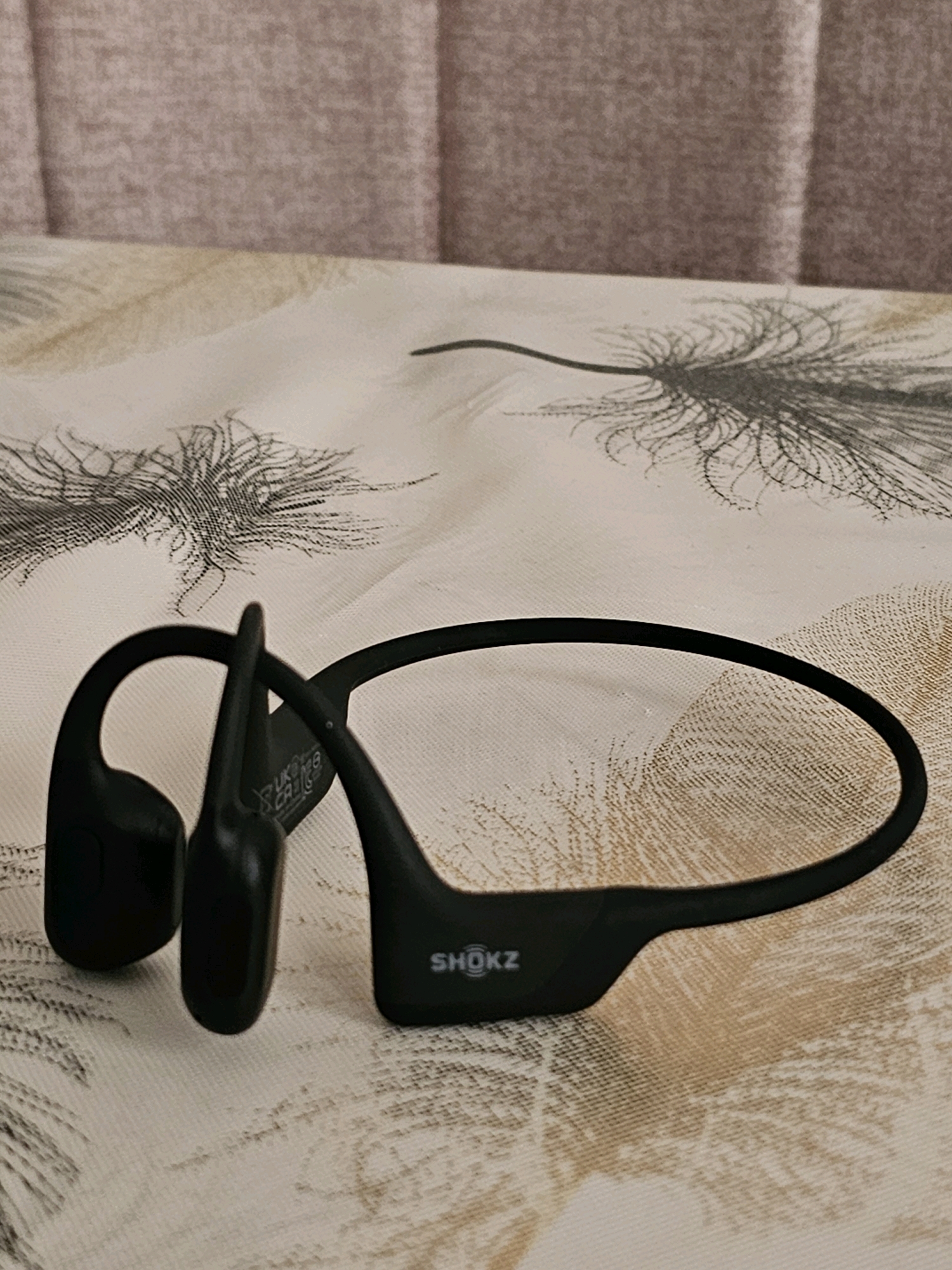 A pair of black Shokz bone conduction headphones is resting on a bed with a light-colored, patterned comforter. The comforter has a design of thin, dark lines that resemble feathers or branches. Behind the comforter, a headboard is visible, upholstered in a textured, light pink fabric. The headphones are positioned in the lower half of the frame, with the left earpiece and part of the band in focus. The right earpiece is further back and slightly out of focus. The brand name "SHOKZ" is clearly visible on the right earpiece. There is no indication of the city of Batna, Algeria, or any other specific geographic location in the image. The scene is static and shows a close-up view of the headphones on the bedding.