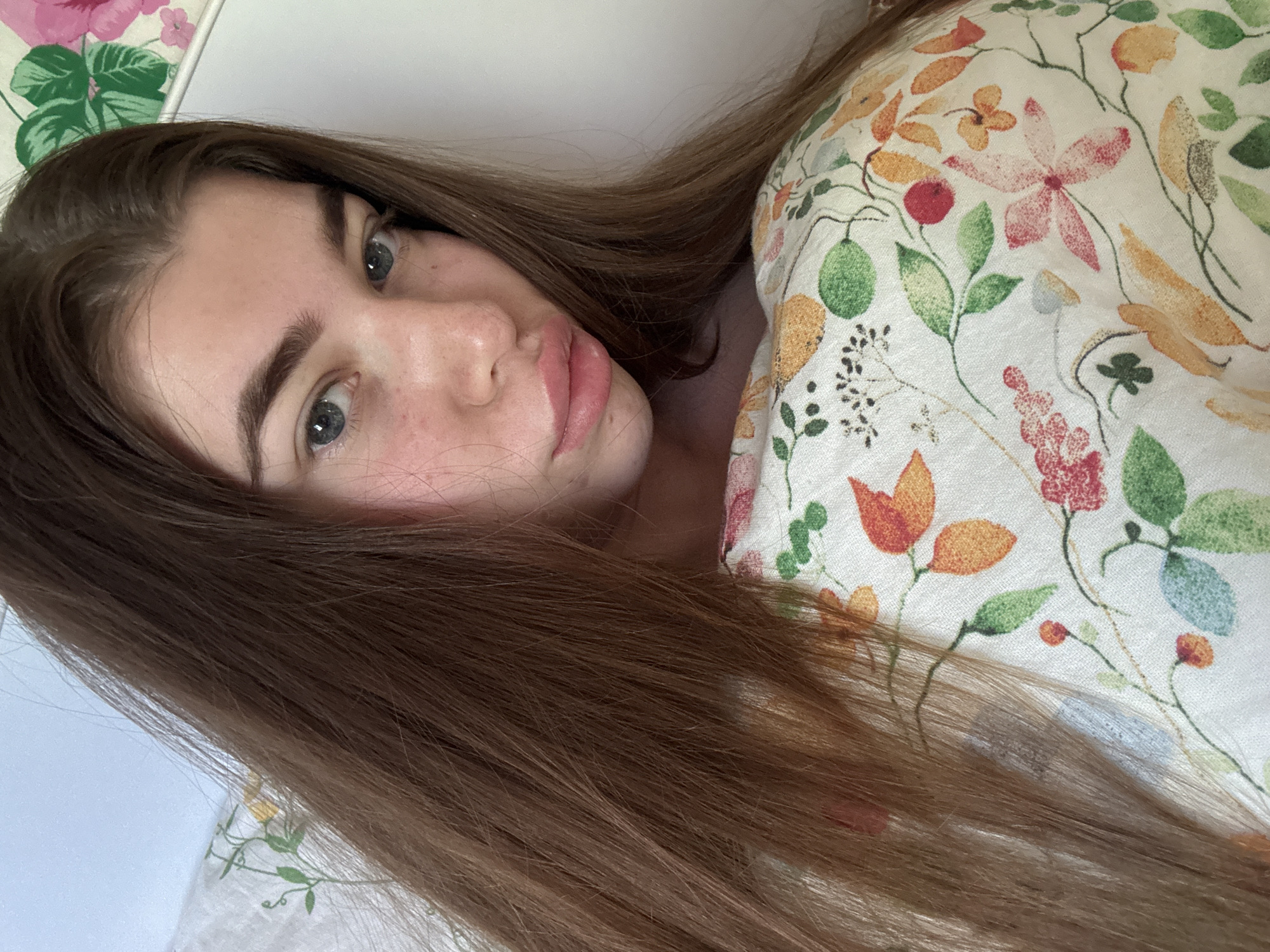 A young woman with light brown hair and blue eyes is lying in bed. She is partially covered by a pillow and bedding with a floral pattern. The room has white walls with a floral design in the top corner. The location is not discernible from the provided image.