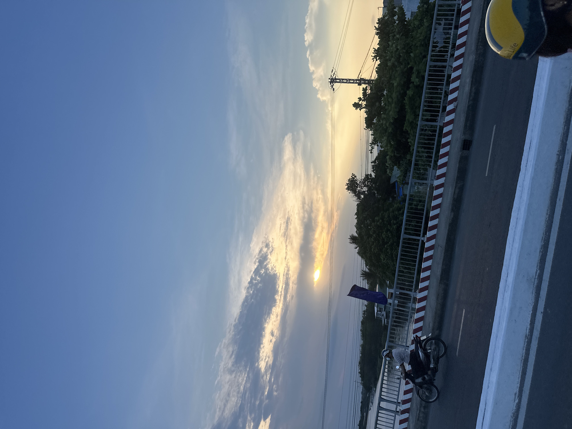 The image captures a sunset scene on a bridge in Nha Trang Ward, Vietnam. A person on a motorcycle is visible on the roadway, adjacent to a guardrail with alternating red and white stripes. Lush green trees and an electrical power pole are present behind the railing. The sky is filled with clouds illuminated by the setting sun, which creates a bright glow and casts shadows across the scene. Part of a yellow helmet is visible in the top right corner.