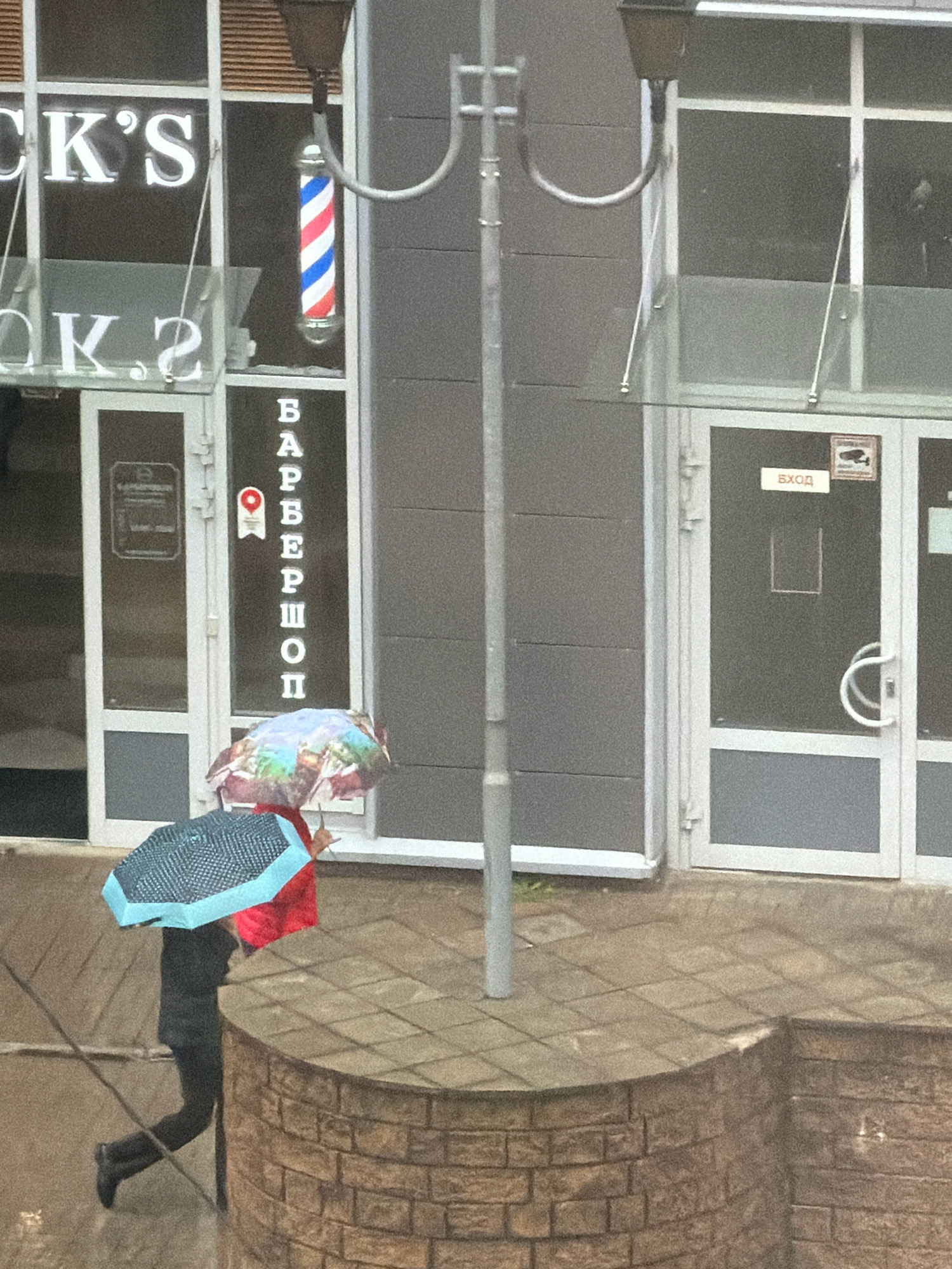 The image shows a street scene in Saint Petersburg, Russia, on a rainy day. A person is walking under two umbrellas, one blue and one red, in front of a building with a barber pole and "BARBERSHOP" written vertically in Cyrillic letters. The building appears to be a commercial establishment, with large glass windows and doors. There is also a sign that reads "ВХОД" (ENTRANCE) on one of the doors. The ground is wet, indicating recent rainfall, and there are puddles visible. The overall atmosphere is somewhat gloomy due to the overcast weather.