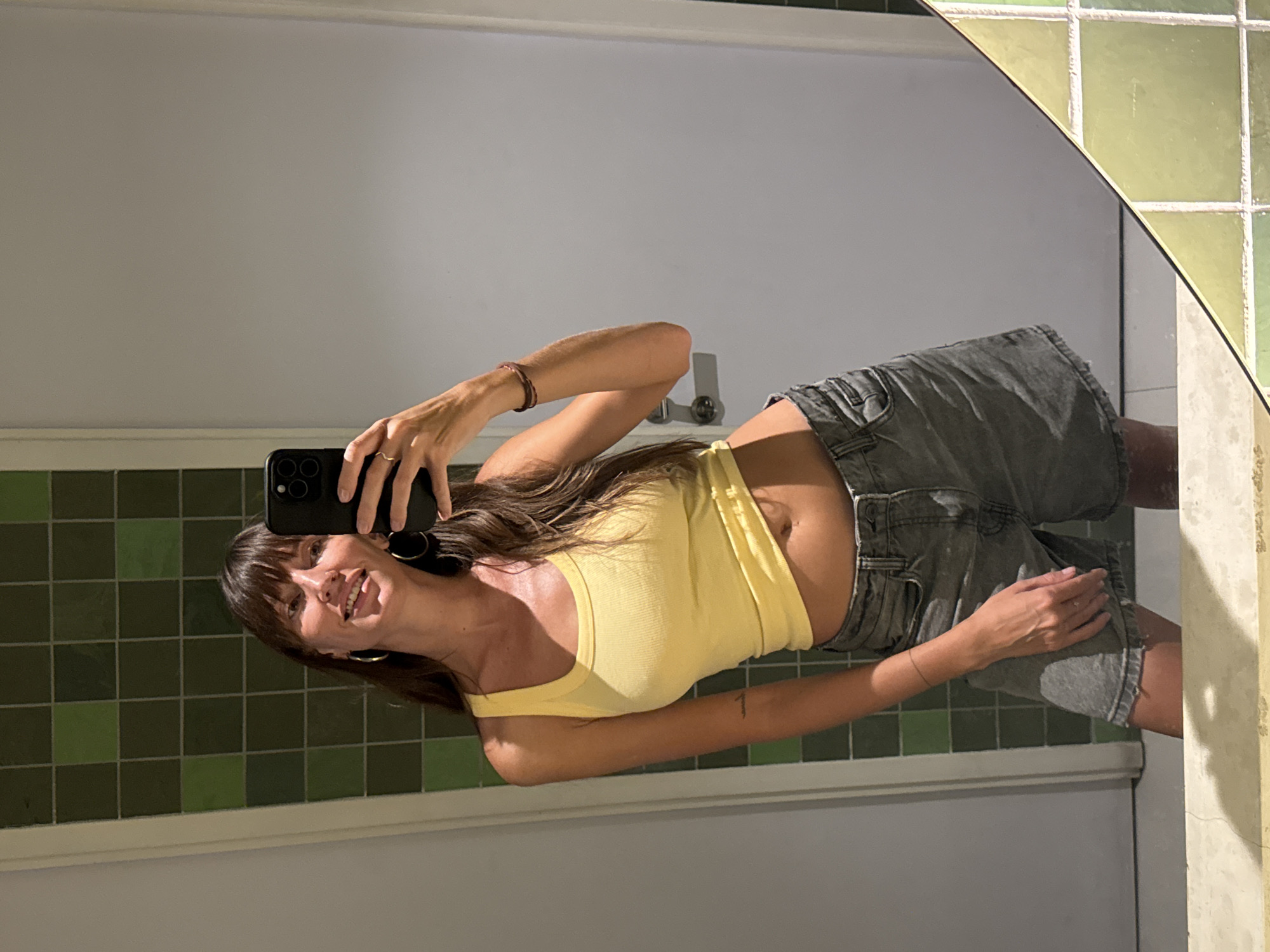 A woman is seen taking a selfie in a bathroom in North Kuta, Indonesia. She is wearing a yellow tank top and gray denim shorts. The bathroom has green tiles and a mirror. The woman is smiling and holding her phone in her left hand. She appears to be in a private residence.