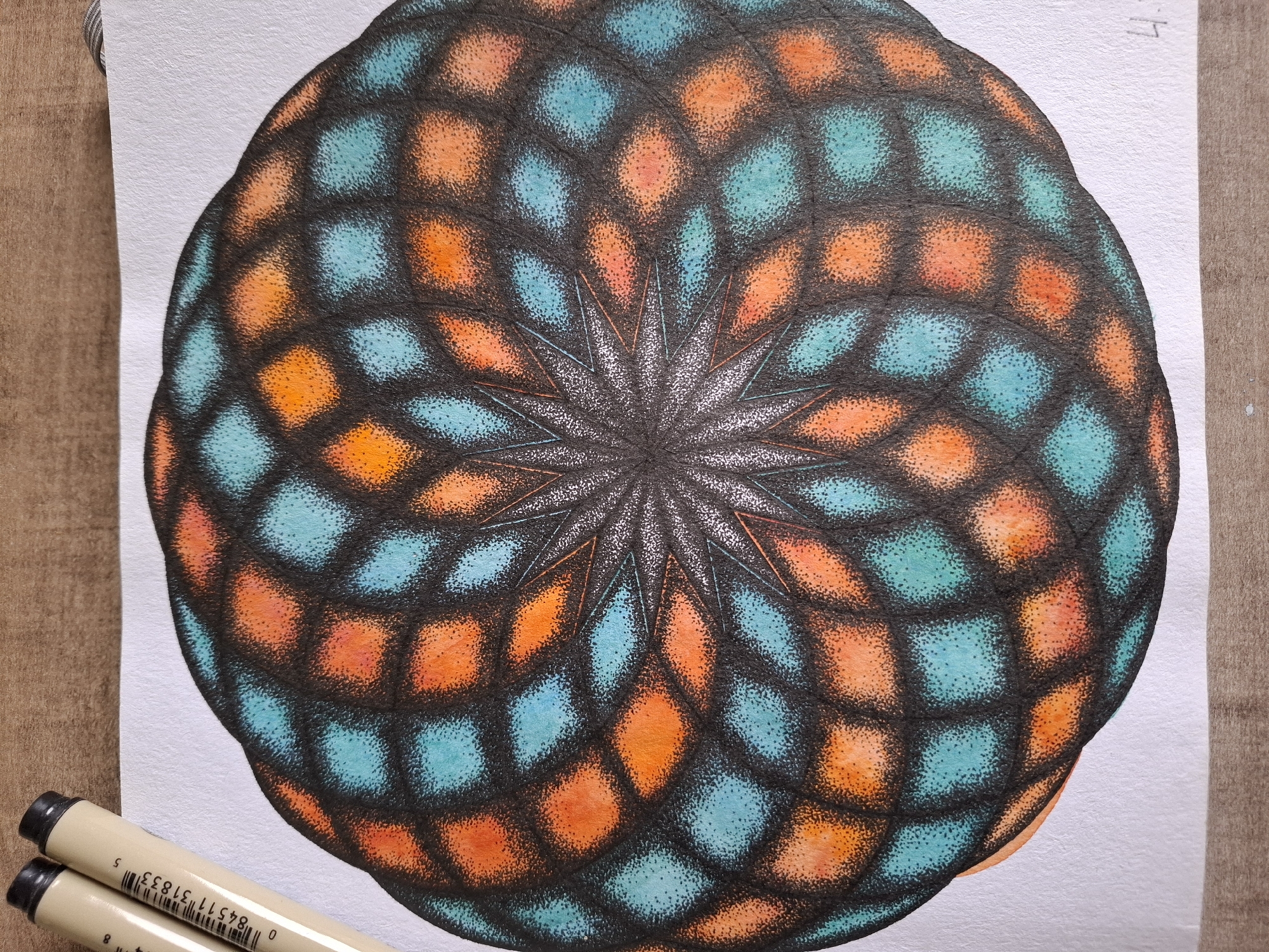 The image shows a close-up, top-down view of a stippled mandala artwork on white paper, placed on a wooden surface. The artwork features a circular design with a starburst pattern in the center, radiating outwards. The design is composed of diamond-shaped segments filled with stippled orange and teal colors, outlined by black stippling. Two black-capped pens, likely technical pens, are positioned in the upper left corner, resting on the paper. The pens have white barrels with black text and markings. The wooden surface appears to be a desk or table. There is no indication of the city of Tyumen, Russia, or any people in the image.