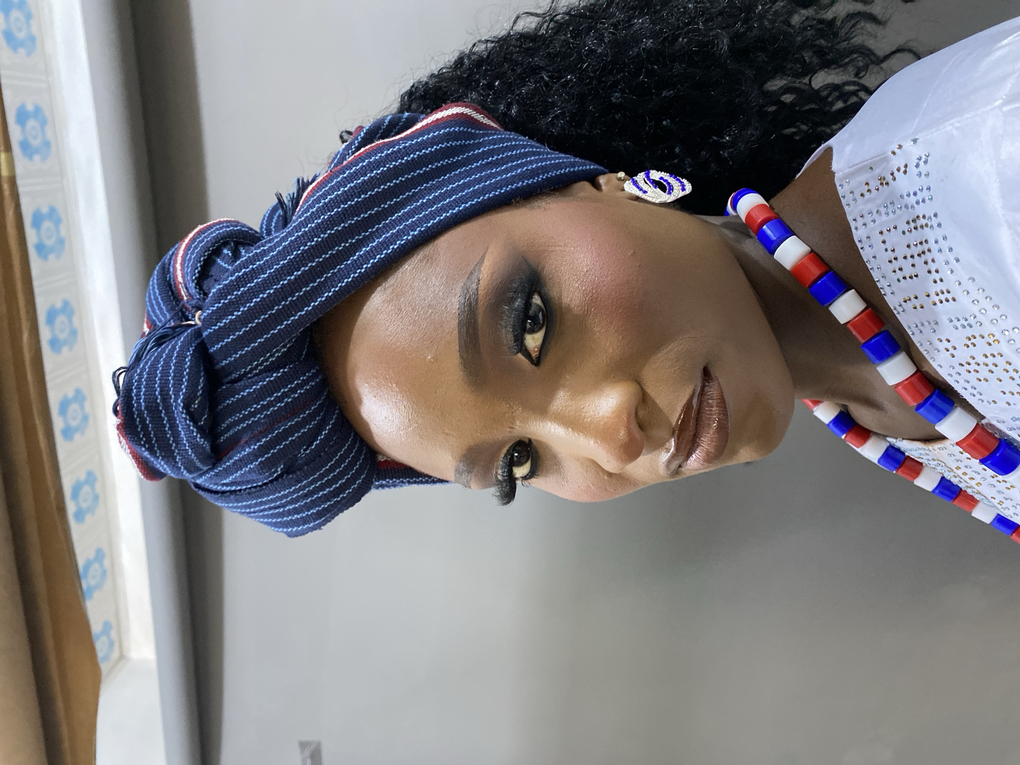 A close-up, head-and-shoulders portrait of a Black woman is shown in a medium shot, positioned at a slight angle to the left. She is wearing a blue and white striped headwrap that is intricately tied, revealing some of her dark, curly hair. Her makeup is dramatic, with dark eyeshadow, defined eyebrows, and glossy, nude-toned lipstick. She is adorned with a white top that has a shimmering, beaded neckline, and is wearing a statement earring on her right ear that is silver and blue. Around her neck, she has a layered necklace made of red, white, and blue beads. The background is a plain, light gray wall. The lighting is soft and appears to be coming from the front. There is no indication of the city of Gura Top, Nigeria, within the image.