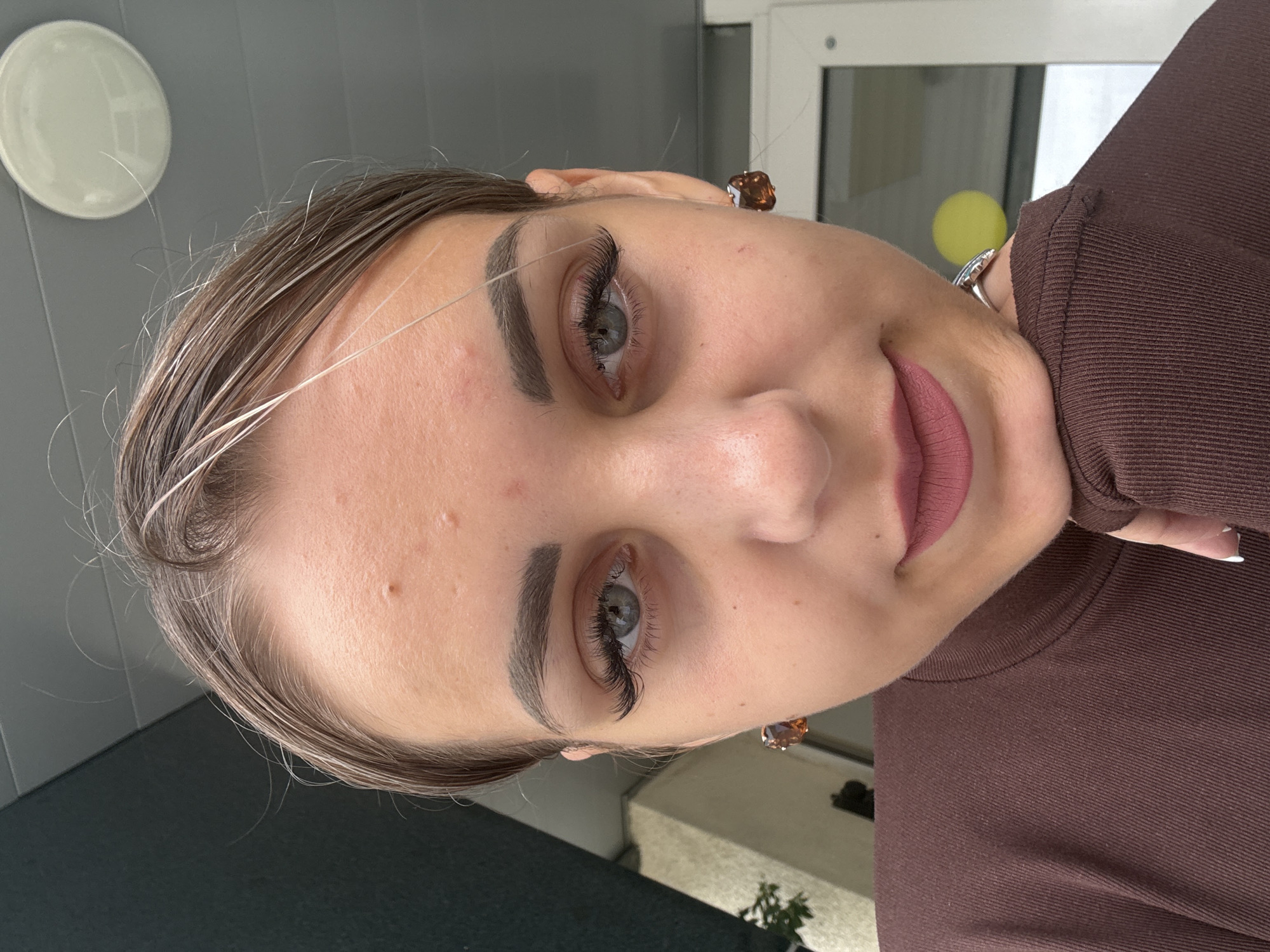 A young woman with blonde hair pulled back, wearing a brown turtleneck, is captured in a close-up, selfie-style portrait. She has notable makeup, including thick false eyelashes and matte pink lipstick. Her gaze is directed towards the camera, and her right hand is resting under her chin, revealing a ring on her index finger. She is wearing large, brown, faceted earrings. The background is out of focus, but appears to be an outdoor setting with a dark, textured wall on the left and a light-colored ceiling with a circular light fixture. A window with a yellow object visible through it is to the right. The scene is not in Yuzhno-Sakhalinsk, Russia, as no identifying features for that location are present.