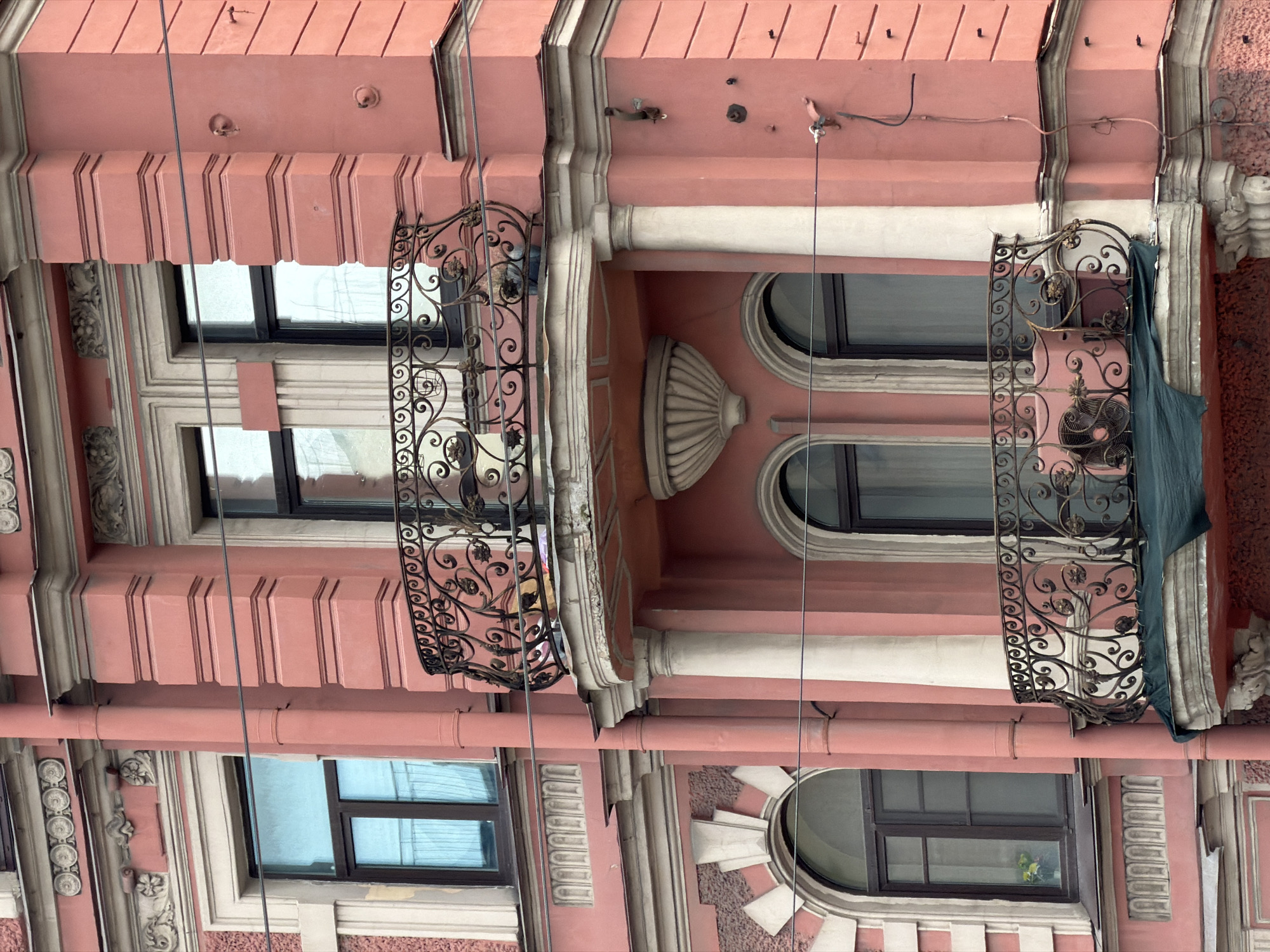 The image shows a close-up view of the ornate facade of a historic building in Saint Petersburg, Russia. The building is painted a salmon pink color and features classical architectural elements, including decorative moldings, columns, and arched windows. There are two balconies visible, both with intricate wrought-iron railings. The upper balcony is supported by columns and has a decorative urn above it. The lower balcony is also adorned with a wrought-iron railing and is partially covered by a green tarp. Wires are strung across the facade. The overall impression is one of decaying grandeur, typical of many buildings in Saint Petersburg's historic center. The time of day appears to be overcast, with diffused lighting. No people are visible in the image.