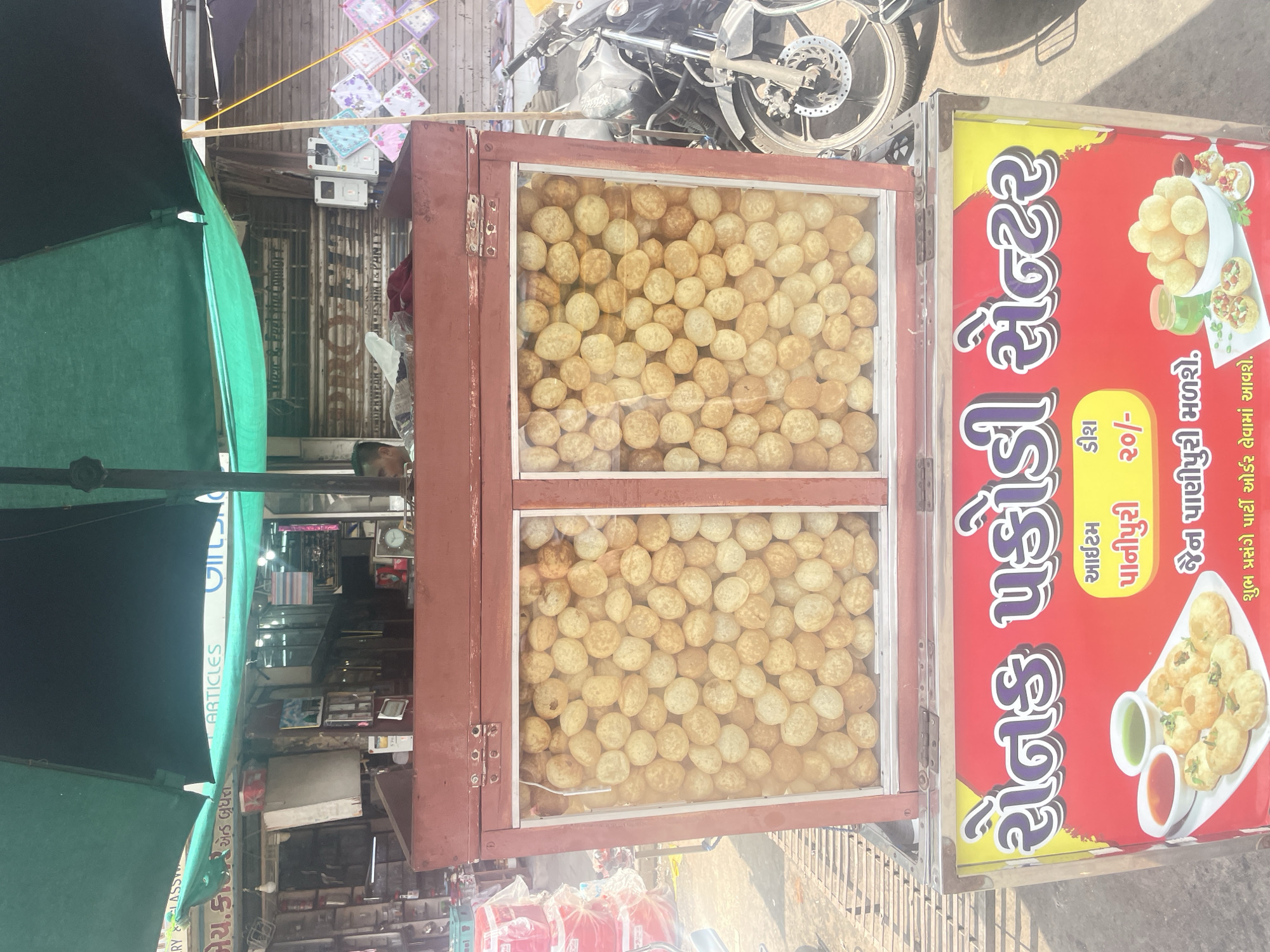 The image shows a street food stall in Ahmedabad, India, specializing in "Panipuri" and "Sev Puri". The stall is covered by a large green and black umbrella. In the foreground, a red and metal display case is filled with panipuri. Below the display case, a sign in Gujarati reads "Sev Puri Center" and advertises "Panipuri" for "₹20/-". The sign also shows an illustration of the food items. To the right of the stall, a motorcycle is parked. In the background, shops and street activity are visible, including a person behind the stall. The overall scene depicts a typical urban street food vending setup.