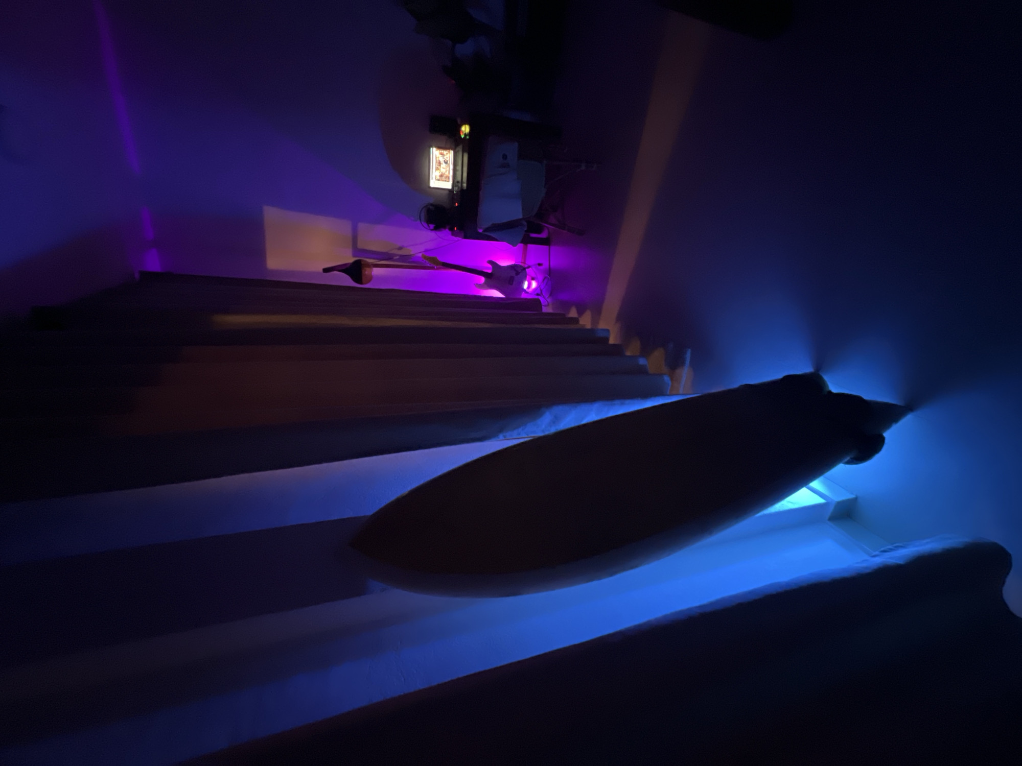 The scene depicts a stairway, viewed from below, illuminated with blue and purple lighting. A long, dark object, possibly a surfboard, rests on the lower steps. At the top of the stairs, equipment including a guitar, music stand and audio setup is illuminated with purple light. The location in Goviyapana, Sri Lanka, is not identifiable from the image.