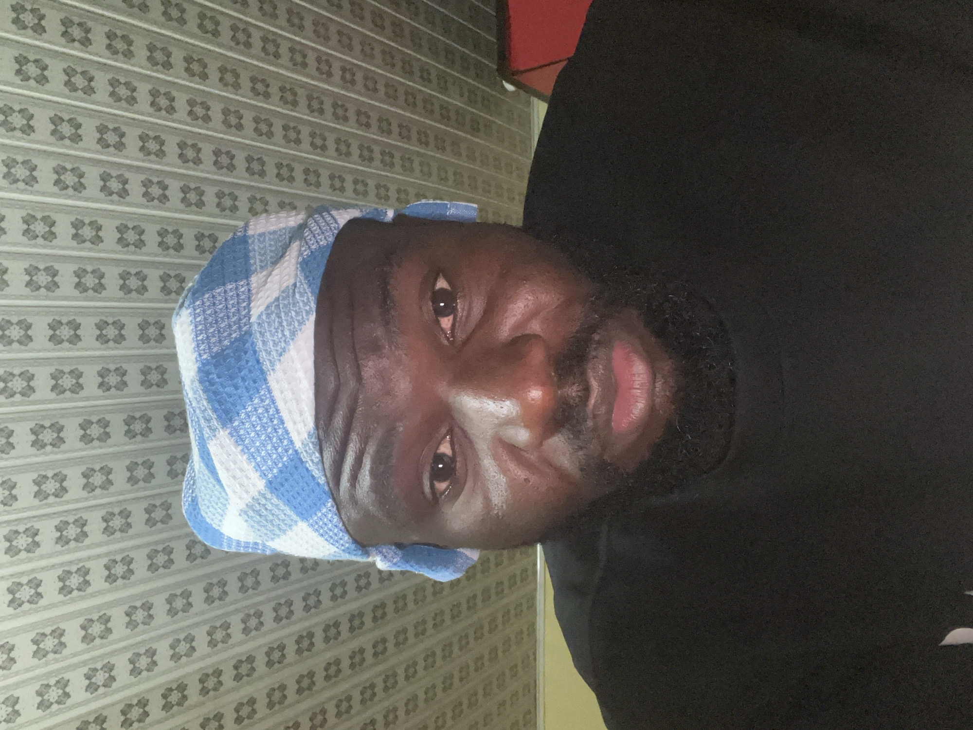A person is seen indoors. The person is wearing a blue and white checkered head covering and a black shirt. The wallpaper behind the person is light grey with a patterned design. The location is Gura Top, Nigeria.
