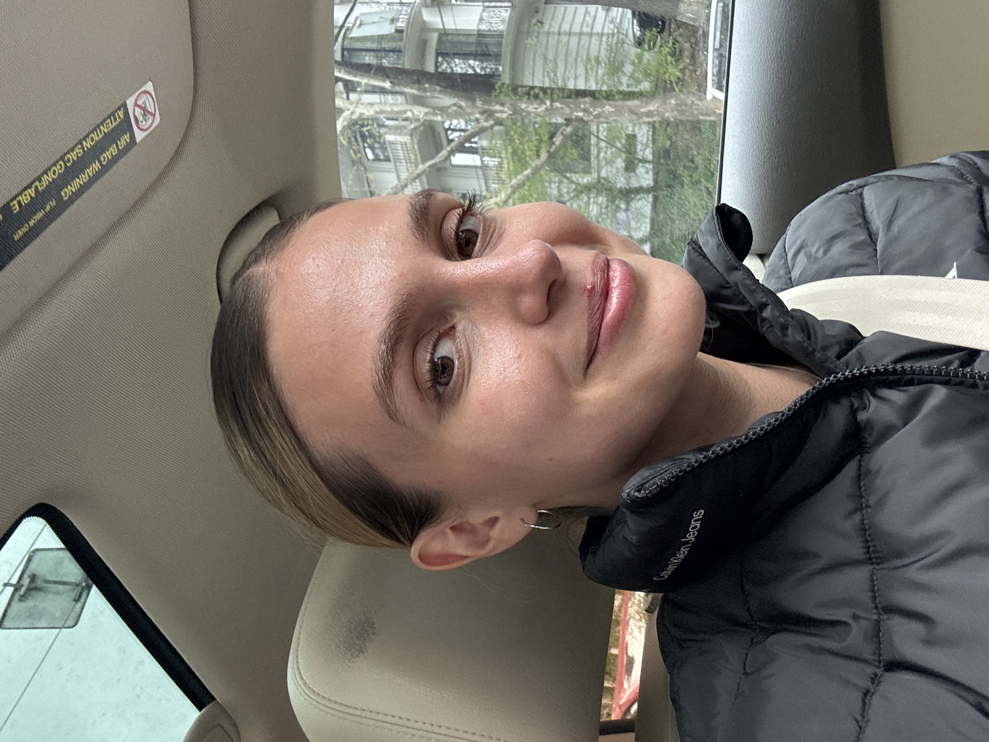 A young woman is captured in a head-and-shoulders portrait taken inside a car, likely in Tbilisi, Georgia, based on the provided context. The image is oriented sideways, with the woman's head resting horizontally. She has light brown hair, styled back, and brown eyes. Her expression is calm, with a slight, soft smile on her lips, which are adorned with a subtle lip gloss. A small blemish is visible on her upper lip. She wears small, silver-colored hoop earrings.
She is dressed in a black puffer jacket, and the text "Calvin Klein Jeans" is visible on the upper left side of the jacket, near her neck or collar. A beige seatbelt crosses her torso. The car's interior features light beige upholstery on the seats and headliner.
Through the car window, visible in the background, are bare tree branches, suggesting a season like late autumn, winter, or early spring. Several buildings are also discernible in the distance. The lighting appears to be natural daylight, possibly from an overcast sky or filtered through trees.
Text is visible on the car's sun visor, which reads "AIR BAG WARNING" and "ATTENTION SAC GONFLABLE," along with a pictogram of a child in a car seat with a diagonal line through it, indicating a safety warning. Below this, smaller text reads "FLIP VISOR OVER."