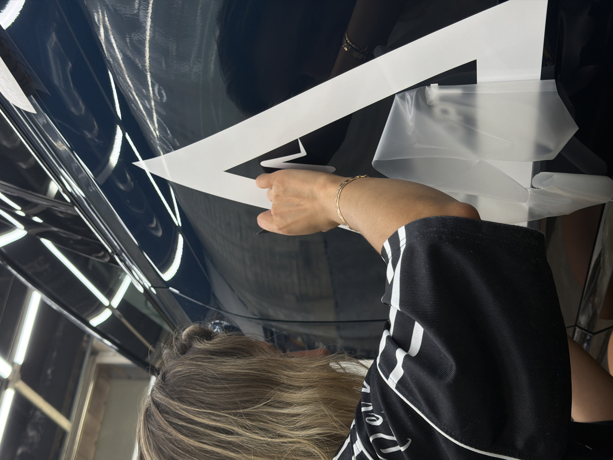 A woman with blonde hair is applying a large white geometric decal to the dark, reflective surface of a car. She is wearing a black shirt with white stripes and a thin gold bracelet on her left wrist. Her right hand holds a tool, possibly a squeegee or a craft knife, and she is carefully working on the decal. The decal appears to be an abstract mountain-like shape or a stylized letter. The car's surface is highly polished and reflects the bright overhead lights of what appears to be an indoor workshop or garage. Part of the decal is still covered in its protective backing, which is peeling away and being held down with clear tape. The background is out of focus but suggests an industrial or commercial setting. The location is not specified.