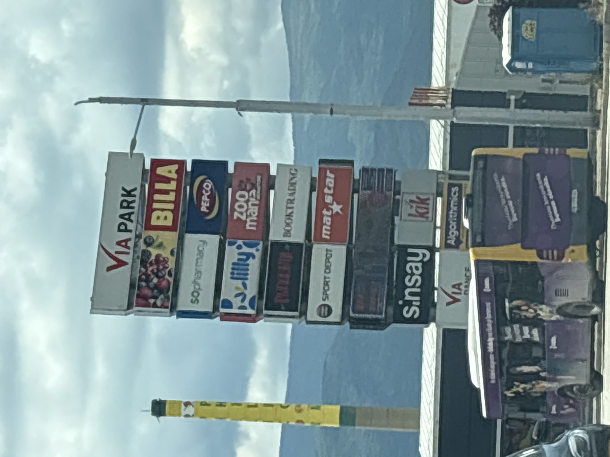 The image shows a tall signpost at Via Park, a shopping center in Plovdiv, Bulgaria. The signpost lists several businesses, including Via Park, BILLA, sopharmacy, PEPCO, Lilly, ZOO mania, BOOKTRADING, SPORT DEPOT, mat star, sinsay, kik, VIA DANCE, and Algorithmics. The sky is cloudy, and there are mountains in the background. The camera is oriented in a sideways position.