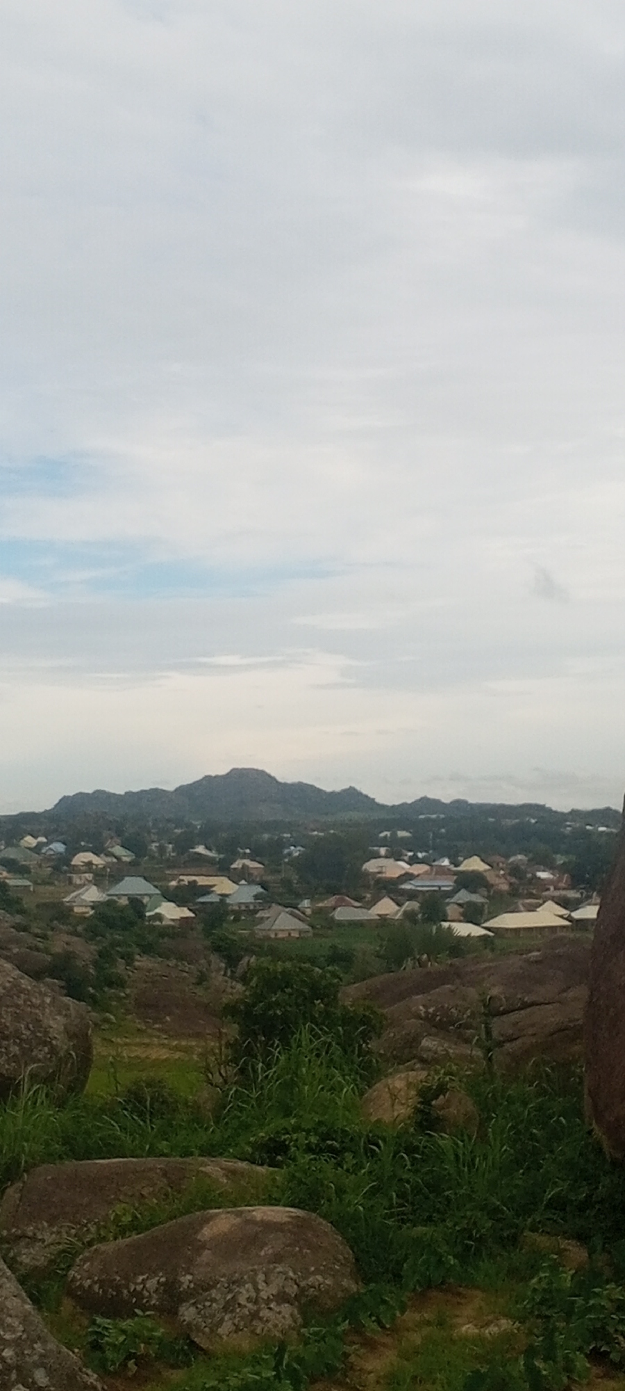 A panoramic view of a densely populated settlement in Ohit, Nigeria, is captured from a rocky, elevated vantage point. The foreground is dominated by large, weathered boulders and lush green grass, indicating a natural, possibly rugged landscape. Beyond this, the midground reveals a sprawling collection of residential buildings, characterized by diverse roofing colors including white, yellow, and blue, suggesting a variety of construction materials and architectural styles. The settlement is nestled at the base of a range of rolling, dark-hued hills that form the backdrop. The sky above is overcast with pale, diffused light, hinting at either dawn, dusk, or a cloudy day. The overall scene depicts a harmonious coexistence of natural terrain and human habitation within a Nigerian rural or semi-urban setting.