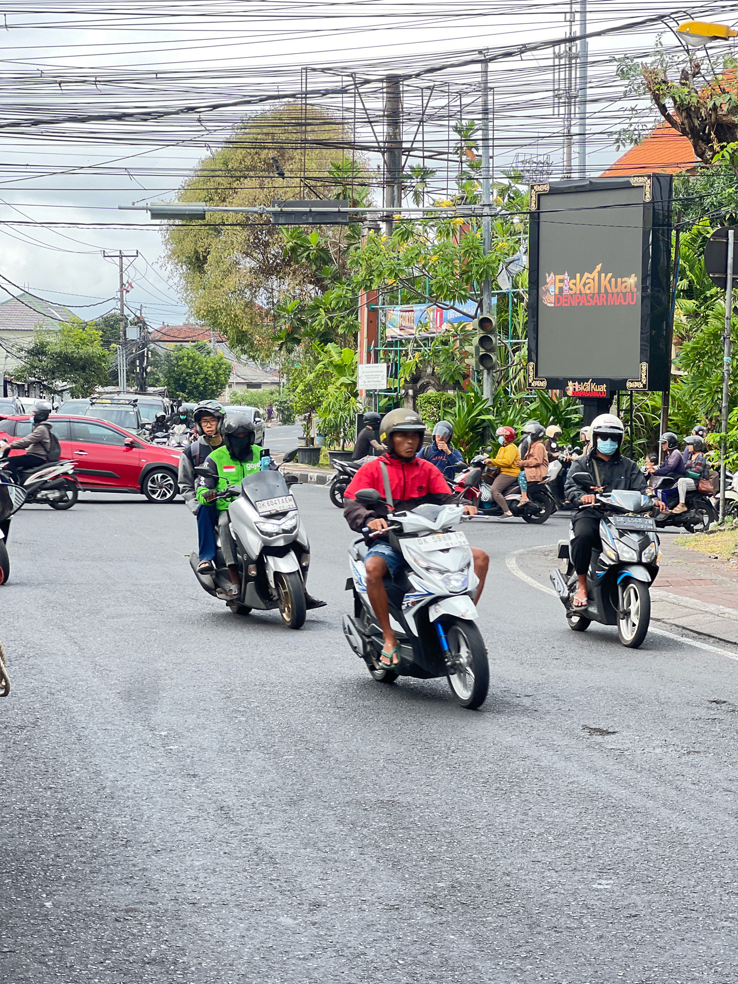 In Denpasar, Indonesia, this image captures a street scene featuring multiple motorcycles with riders wearing helmets and some with face masks. The motorcycles are on a paved road, and traffic appears to be moderate. On the left side of the road, a red car is visible. In the background, there are trees, buildings with orange roofs, and power lines crisscrossing overhead. A large digital billboard displays text and graphics, with "Fiskal Kuat" and "DENPASAR MAJU" prominently featured. The sky is overcast, suggesting a cloudy day.
