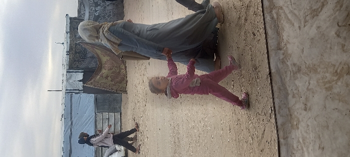 The image depicts a scene in Khan Yunis, Palestinian Territory, featuring four individuals on a dirt path with makeshift shelters in the background under an overcast sky. In the foreground, a young girl, estimated to be between 3 and 5 years old, wearing a pink hooded tracksuit and pink shoes, walks towards the right while looking slightly towards the camera. Her left hand holds the right hand of an adult woman walking beside her, also towards the right. The woman wears a light blue full-length garment and a light-colored head covering, obscuring her face from the camera. In the mid-ground, two additional individuals walk from left to right. One wears a light-colored top and dark pants, and the other is largely obscured but appears to wear dark clothing. The ground is unpaved, consisting of light-colored dirt or sand, with a concrete or paved edge visible at the very bottom of the frame. The background features several rudimentary structures, likely temporary shelters, constructed from materials including white tarpaulins, corrugated metal sheets, and patterned fabric blankets. A dark, thin pole stands vertically behind these structures.