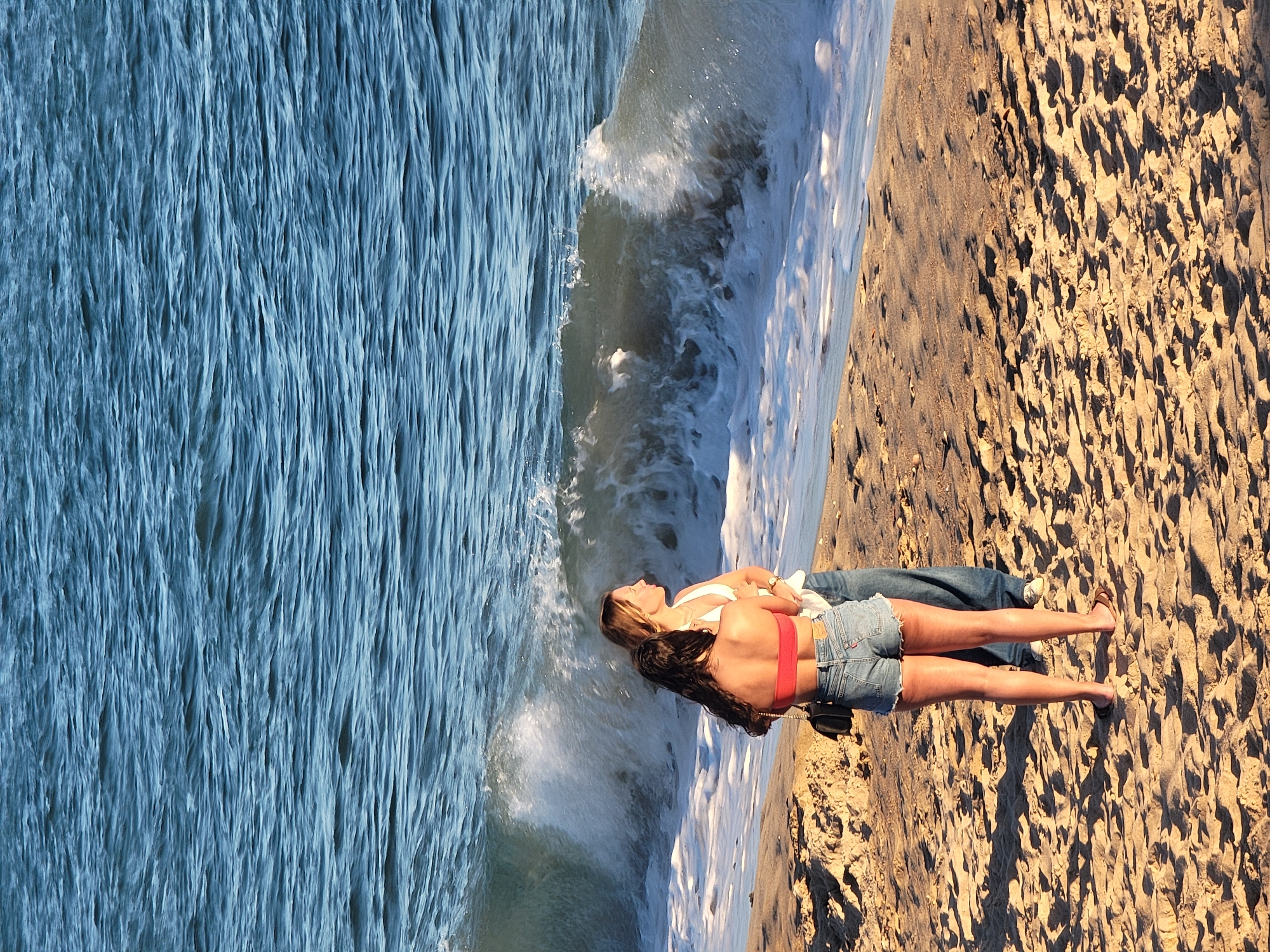 Two individuals stand on a sandy beach in Quarteira, Portugal, facing the ocean as a wave breaks. The person in the foreground, wearing a red bikini top and denim shorts, has their back to the viewer, with a black purse slung over their shoulder. The second individual, to their right and slightly behind, wears a white top and blue jeans and is facing the ocean. The water is a deep blue, with white foam from the breaking wave washing onto the shore. The beach is covered in coarse sand, with shadows cast by an unseen light source, likely the sun. The scene captures a moment of observation of the natural environment.