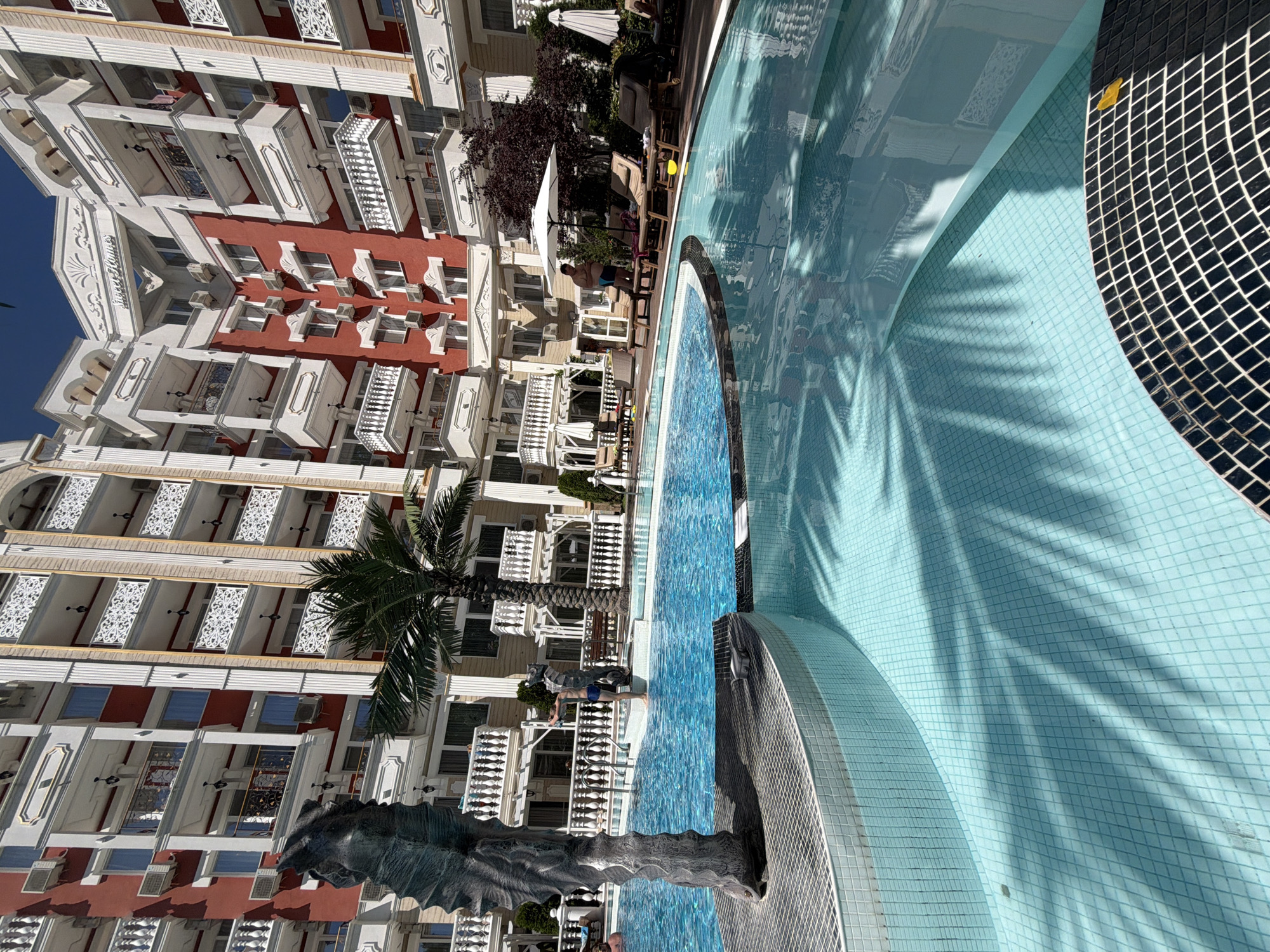 This image depicts an outdoor swimming pool area in front of a large, multi-story hotel building. The hotel has a distinctive architectural style with red and white facades, ornate balconies with white railings, and arched window frames. Palm trees are visible around the pool, and lounge chairs and umbrellas are set up on the patio. The swimming pool is a prominent feature, with clear blue water and a tiled bottom. Shadows of palm fronds are cast across the water's surface. In the foreground, a dark, abstract sculpture is partially visible to the left of the pool. A young boy in blue swim trunks stands near the edge of the pool, facing away from the camera, with a seahorse-shaped sculpture beside him. Another person is partially visible in the background near the pool chairs. The sky is clear and blue, suggesting a sunny day. The hotel appears to be named "Sweet Homes" based on signage at the top of the building. This scene is located in the city of Nesebar, Bulgaria.