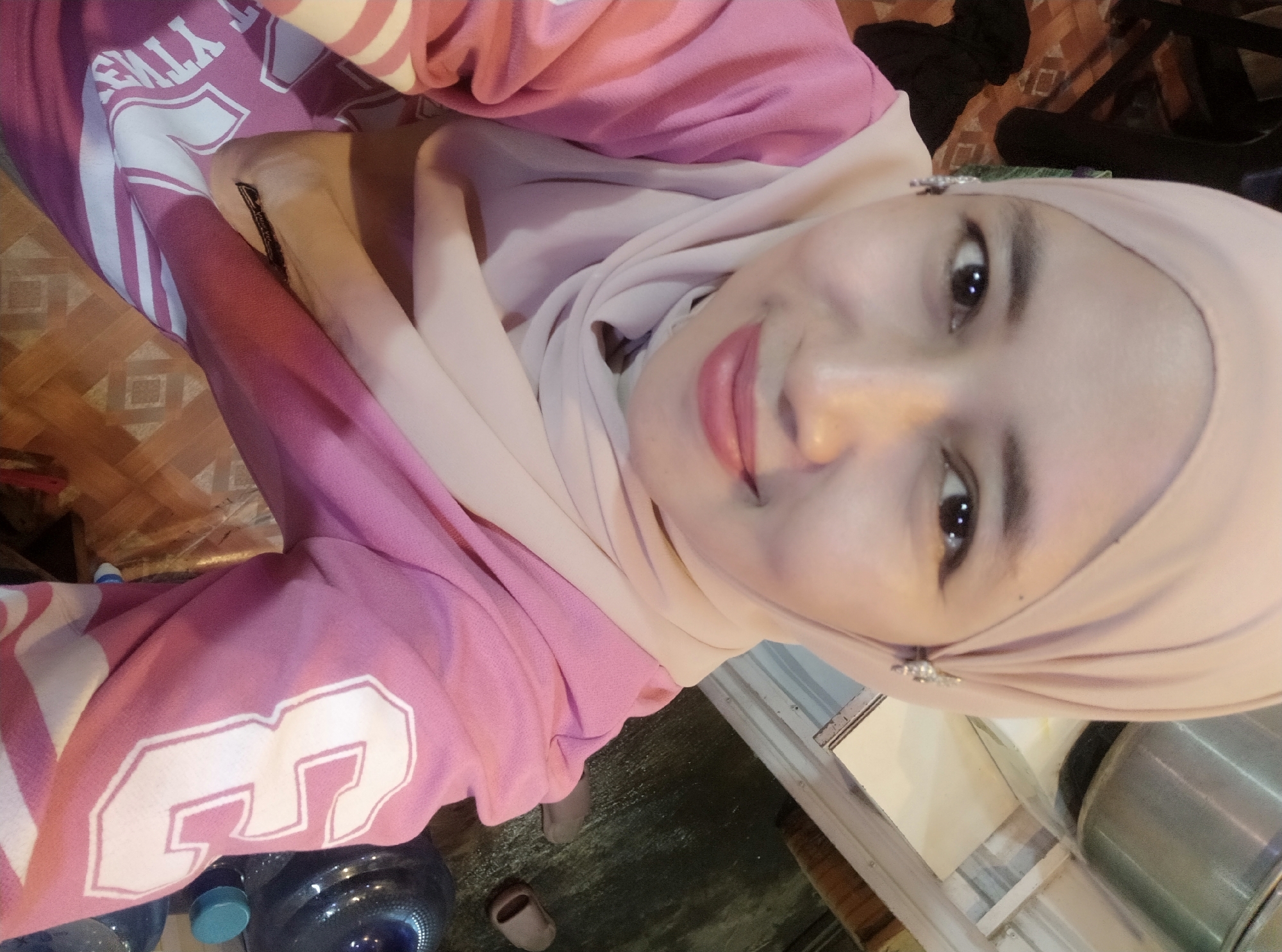 A young woman is shown from the chest up, taking a selfie. She is wearing a light pink hijab and a pink long-sleeved shirt with white stripes. She has light skin and is smiling. She is accessorized with small, sparkling earrings. The background is blurred and appears to be indoors, with some shelving and what looks like a large container. Based on the clothing and style, the image is likely from Subah, Indonesia.
