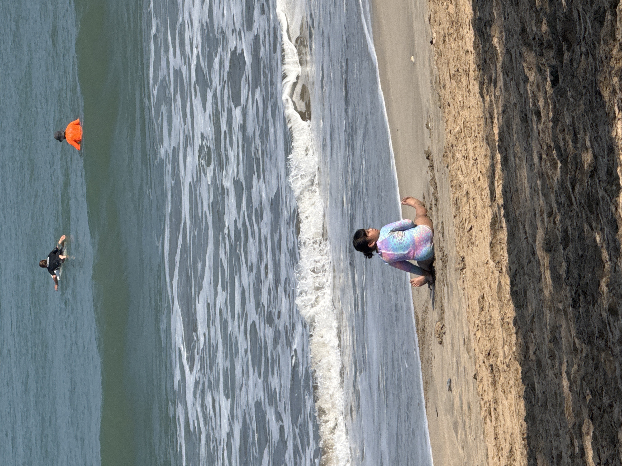 The image depicts a beach scene with two individuals in the water and one on the sand. In the water, a person is wearing an orange top and a dark hat, appearing to be partially submerged. Further to the left, another individual in a black wetsuit is in the water, seemingly engaged in an activity like surfing or bodyboarding, with their arms outstretched.
On the sandy shore, a person is sitting with their legs tucked underneath them, looking out towards the water. They are wearing a colorful long-sleeved swimsuit with a tie-dye pattern. The waves are breaking on the shore, creating white foam. The sand appears dry and textured, with some rocks or debris scattered on it. The background shows a steep, rocky cliff face.
The scene suggests a sunny day, likely late afternoon given the shadows cast by the cliff, and a moderate amount of surf. The individuals appear to be enjoying recreational activities at the beach. The location context provided indicates Kuta, Indonesia, a well-known surf destination. There is no visible text in the image.