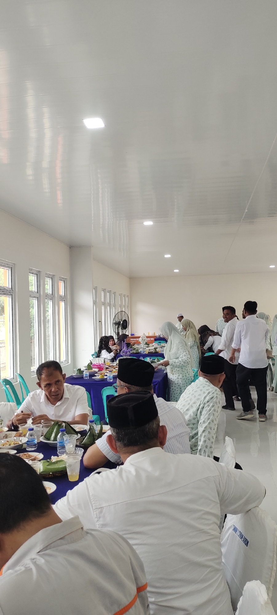 In a well-lit indoor hall, possibly a community center or dining area, a group of people are gathered for a meal or event. The room features high ceilings with modern lighting fixtures and large windows along one wall, offering glimpses of greenery outside. The atmosphere appears to be formal, with many attendees dressed in white attire and peci hats, suggesting a religious or cultural occasion.
In the foreground, several men are seated at tables covered with blue tablecloths. One man in a white shirt is speaking, gesturing with his hands, while others listen or look towards the food laid out before them. The tables are set with plates of food, wrapped packets, and drinks in plastic cups and bottles.
In the background, more individuals are standing and mingling. Some women in headscarves are also present, including one wearing a light-colored, patterned outfit. A large black oscillating fan is visible near the windows, likely for air circulation. A sign with text, partially obscured, is mounted on the wall. The floor is a light-colored tile. The overall impression is of an organized gathering in a clean, functional space within Meucat, Indonesia.