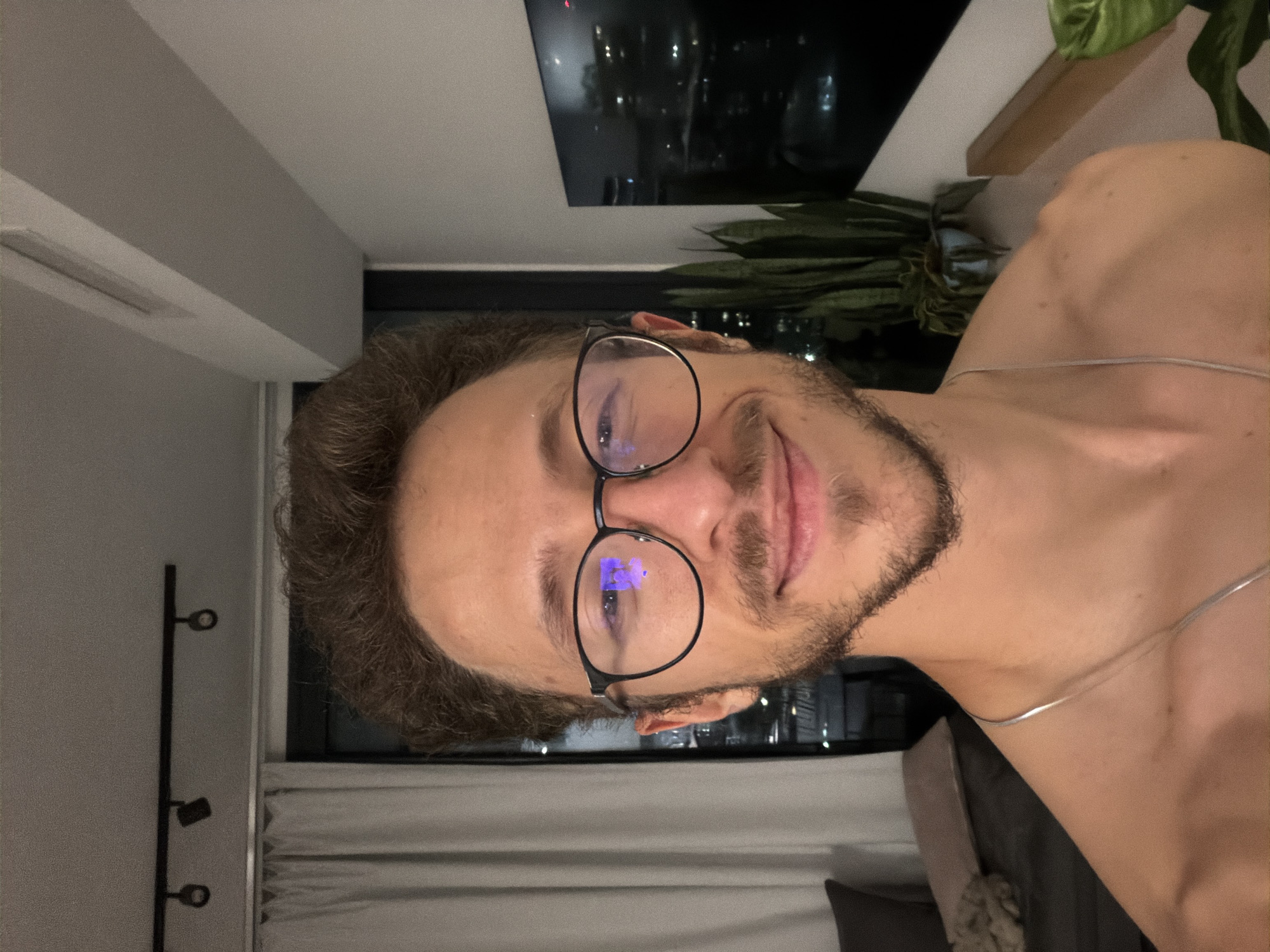 An indoor, close-up portrait of a smiling man is presented, rotated 90 degrees clockwise. The man has curly brown hair, a neatly trimmed beard, and wears round, black-framed glasses that reflect blue light. He is shirtless, revealing his upper chest and shoulder, and a thin silver chain rests around his neck and shoulder. He looks directly at the camera with a relaxed and pleasant expression.
The background suggests an evening or nighttime setting. Through a window behind him, out-of-focus city lights are visible, indicating an urban environment. To the right, a large green plant with broad leaves is discernible, partially obscured. Above the plant, a dark, flat surface or frame is visible. On the left side of the frame, there's a wall with a slender, dark, vertical fixture featuring circular elements, possibly a coat stand or a modern light fixture. Below that, a white window blind or curtain is partially visible. The indoor lighting is subdued, highlighting the man's face and creating a calm atmosphere. No visible text is present in the image.
