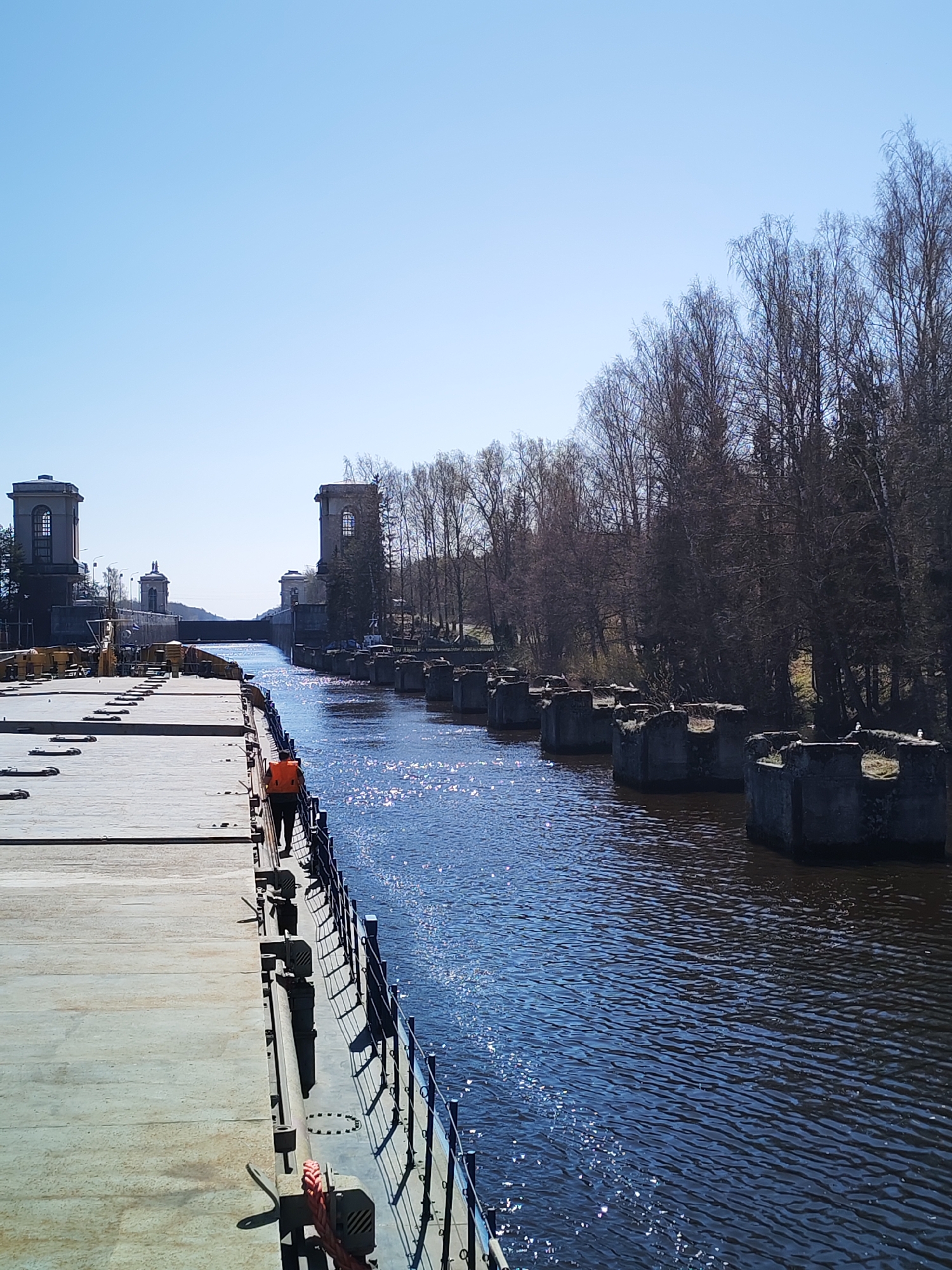 The image shows a canal in an unknown city. The canal is lined by a concrete pier on the left and a row of old, concrete structures in the water on the right, beyond which is a line of trees. A person wearing an orange vest walks along the pier. In the distance are buildings with towers alongside the canal. The sky is clear and blue.