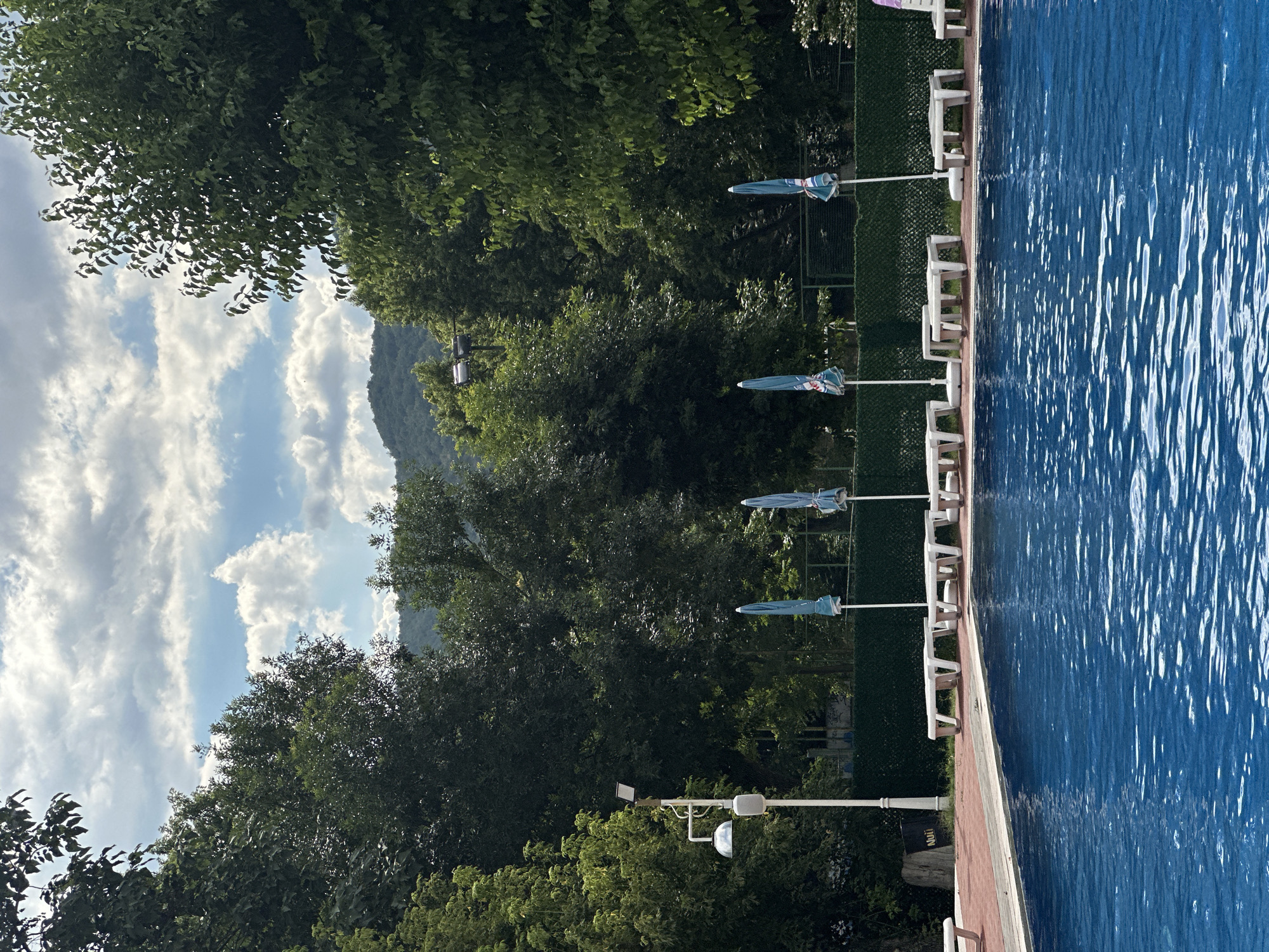 The image depicts a sunny day at an outdoor swimming pool located in Simitli, Bulgaria. The pool is filled with clear blue water, with ripples reflecting the sky and surrounding trees. Along the edge of the pool, there is a red tiled walkway and several white lounge chairs, some of which are empty. A few closed blue umbrellas are set up behind the lounge chairs. In the background, a dense line of lush green trees forms a natural backdrop, with a slight incline of a hill visible behind them. The sky is partly cloudy, with fluffy white clouds against a bright blue expanse. A single lamppost stands to the left of the pool area, with a modern light fixture and a curved shower head. The overall impression is one of a serene and well-maintained recreational area.