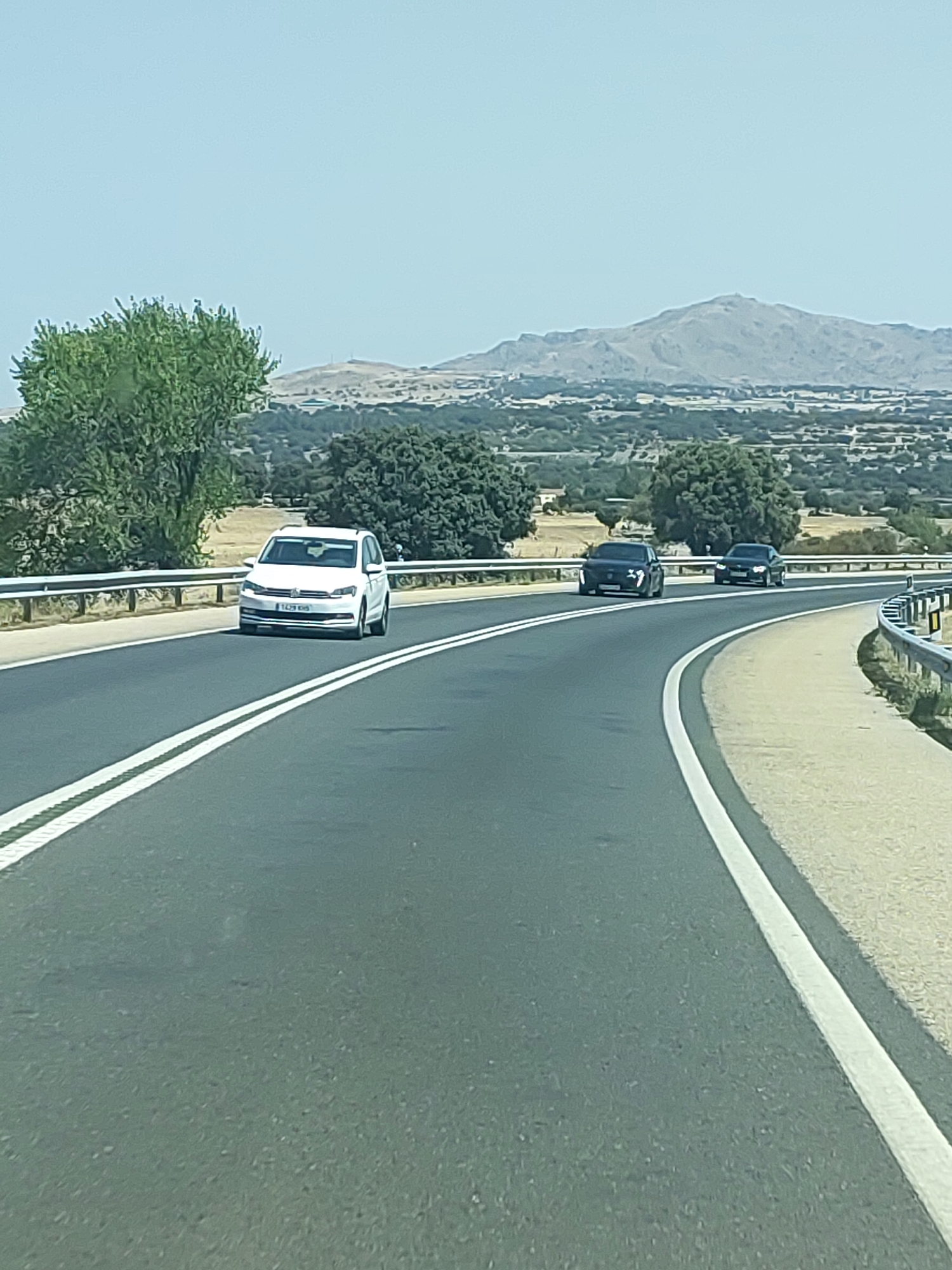 The image shows a view from a vehicle traveling on a highway in Manzanares el Real, Spain. The highway has two lanes in the direction of travel, marked by solid white lines. In the foreground, the asphalt road is visible with lane markings. On the left side of the road, a guardrail separates the highway from the surrounding landscape. On the right side, the road curves to the right, with a sandy shoulder and a similar guardrail.
In the middle ground, three cars are traveling on the highway. The closest car, on the left lane, is a white Volkswagen Touran. To its right, in the right lane, is a dark-colored sedan, possibly a BMW. Further ahead in the right lane is another dark-colored sedan.
The background of the image shows a hilly, rural landscape with scattered trees and vegetation. In the far distance, a mountain range is visible under a clear, bright sky. The overall scene depicts a typical highway environment in a rural Spanish setting.