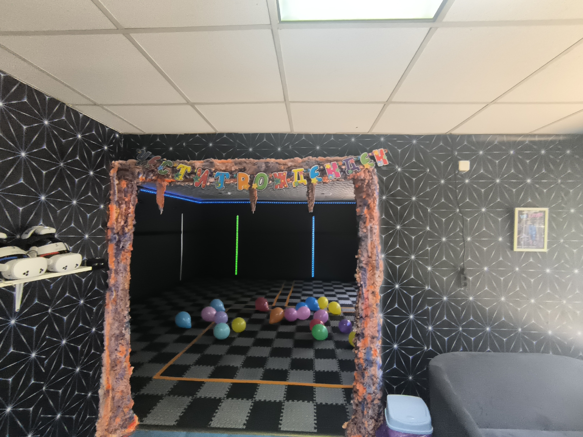 The image captures an indoor entertainment or party room, likely a virtual reality arcade, located in Trastenik, Bulgaria. The walls are covered in dark wallpaper featuring a repetitive geometric pattern of white lines. The ceiling is a standard white drop ceiling with fluorescent lighting panels.
A prominent archway, decorated with fuzzy, multi-colored material in shades of orange, brown, and purple, frames the entrance to an inner, darker play area. A colorful banner hangs across the top of this archway, displaying Cyrillic text that reads "С ДНЁМ РОЖДЕНИЯ," which translates to "Happy Birthday."
Inside the archway, the floor is covered with black and grey interlocking mats, forming a checkered pattern, with orange tape marking specific areas. Numerous partially deflated balloons in various colors, including blue, pink, yellow, green, orange, and purple, are scattered across this floor. Two vertical strips of colored lights, one green and one blue, are visible on the far wall of the inner space.
To the left of the archway, a white shelf holds several white virtual reality headsets. To the right, a dark grey sofa is partially visible, alongside a small, light blue lidded trash can. A framed picture with a yellow border hangs on the wall above the sofa. The scene suggests a recently held birthday celebration, possibly at a VR gaming venue. No individuals are present in the image.