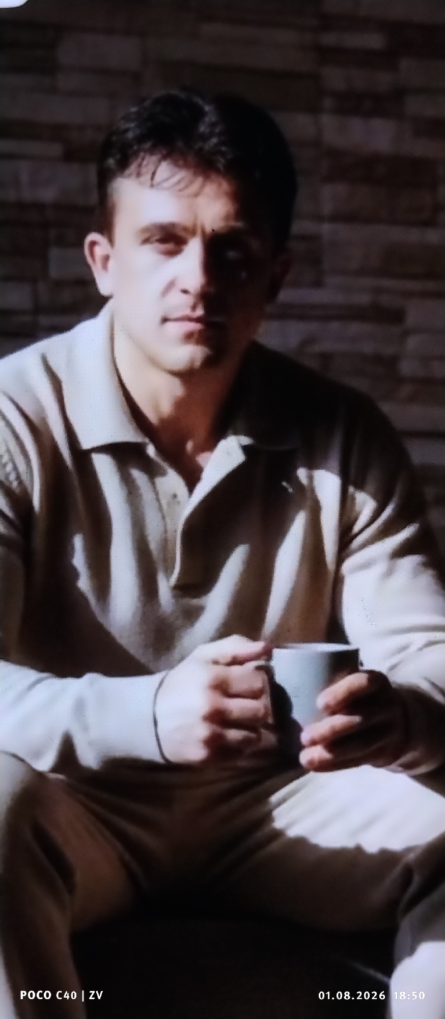A medium shot shows a man sitting in an indoor environment, holding a mug. He is wearing a light-colored collared shirt. His face is partially shadowed, with his eyes visible. The background appears to be a textured wall. The bottom of the image displays a watermark "POCO C40 | ZV" on the left and a date and time stamp "01.08.2026 18:50" on the right. The location is specified as Лубянки, Russia.