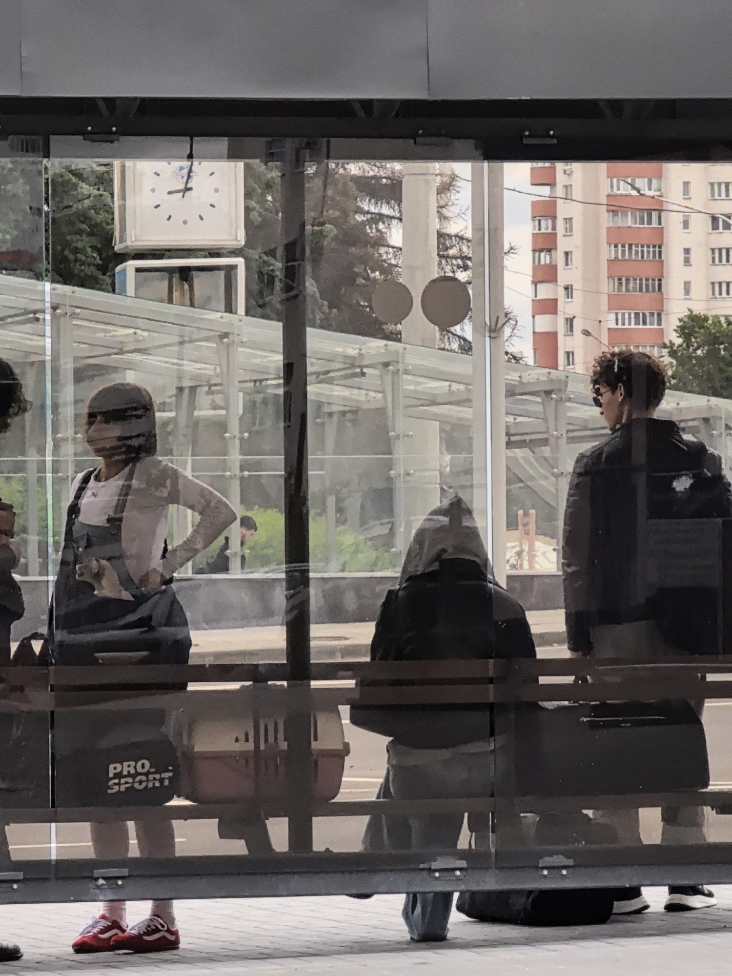 The image depicts a group of people at what appears to be a bus stop or sheltered waiting area. A clock is visible in the background, suggesting a public transportation setting. In the foreground, a person wearing overalls and a t-shirt is standing, with a pet carrier in front of them. To their right, another person is seated and facing away from the camera. Further to the right, a third person wearing a dark jacket and sunglasses is also standing and looking away. The environment appears to be urban, with a building visible in the distance. The people are casually dressed and appear to be waiting.