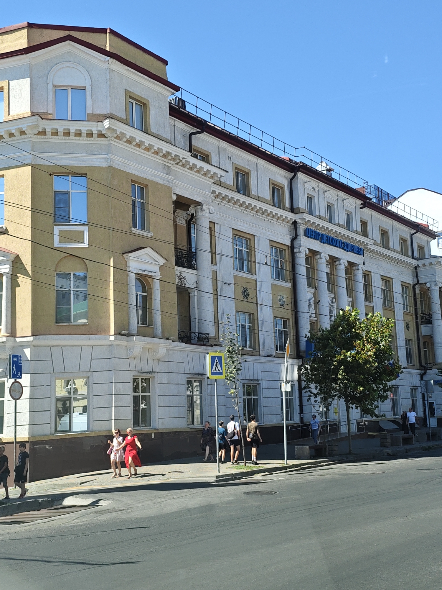 The image depicts a sunny daytime scene on a city street in Rostov-on-Don, Russia, specifically in front of a large, multi-story building with a light yellow and white facade. The building exhibits classical architectural elements, including columns, decorative moldings, and arched windows. Blue lettering is visible across the upper portion of the building, reading "ЦЕНТР ДЕТСКОГО ЗДОРОВЬЯ," which translates to "Center for Children's Health."
Several individuals are present on the sidewalk and crossing the street. On the sidewalk in the foreground, a group of four young people, two males and two females, are walking from left to right. Further down the sidewalk, two women in dresses are walking together, one in a red dress and the other in a lighter pink dress. To the right, a group of several people are walking or standing near the building entrance. A few individuals are also visible further down the street.
Street furniture includes a pedestrian crossing sign with a yellow diamond shape and a pedestrian symbol, as well as a blue and white sign indicating a bus stop with a pedestrian icon. A mature tree with green foliage stands on the sidewalk in front of the building, partially obscuring the middle section. Power lines are strung across the scene, indicating an urban environment. The road in the foreground is asphalt, with a pedestrian crossing marked by white lines. The overall impression is of a typical urban streetscape on a clear day.