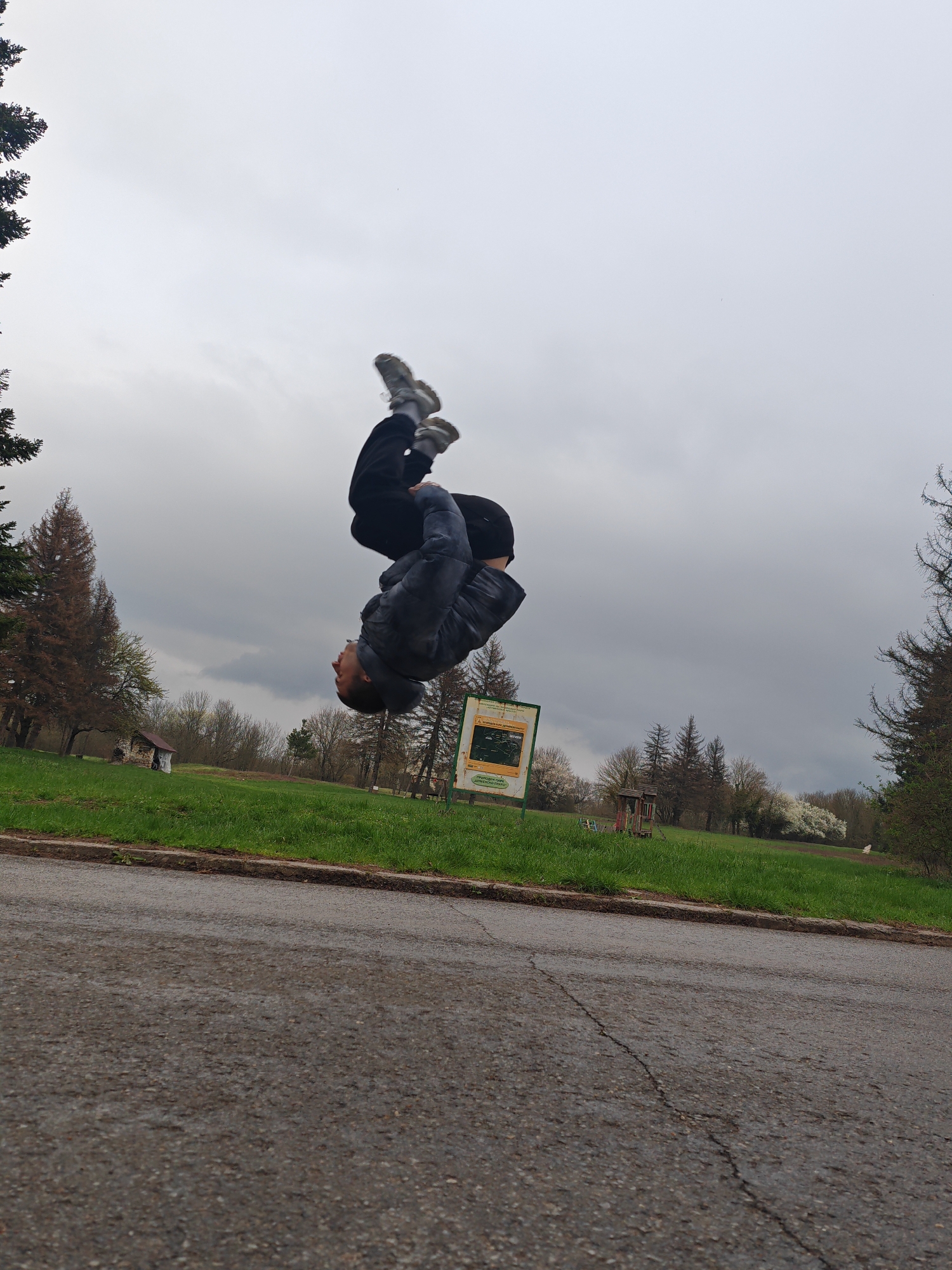 A man in mid-air, performing a backflip on a paved surface. He wears dark pants, a dark jacket, and light-colored shoes. The location appears to be a park, possibly near a road, with grass, trees, and a cloudy sky. In the background are green trees, some with brown foliage, green grass, and a wooden playhouse, possibly located in Shumen, Bulgaria. There is also a information/ park map sign. The pavement has a crack running across it.
